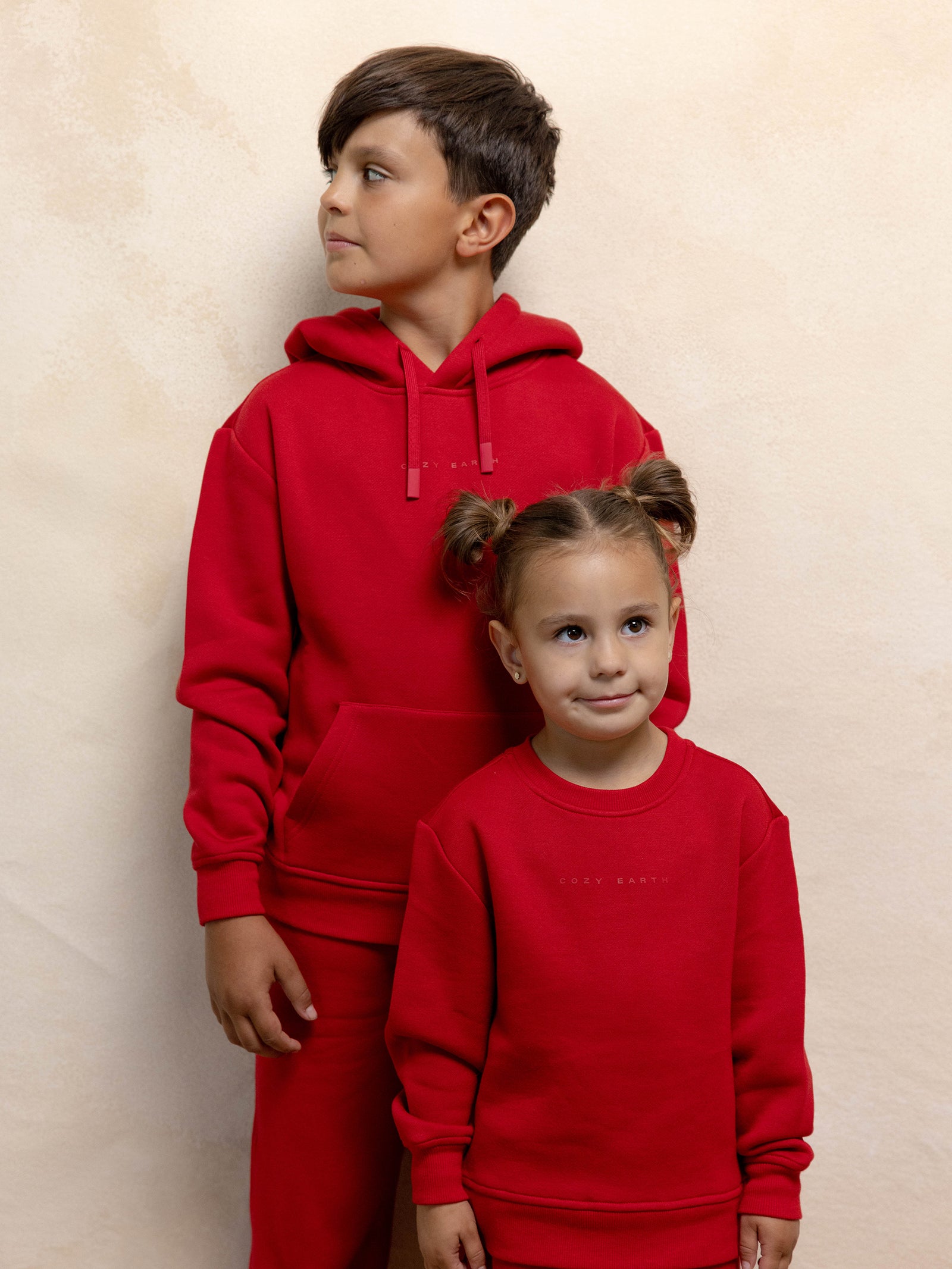 Two children in matching red Kid's CityScape Crewnecks by Cozy Earth stand in front of a beige wall. The older child, a boy, looks to the side while the younger child, a girl with pigtails, smiles at the camera.