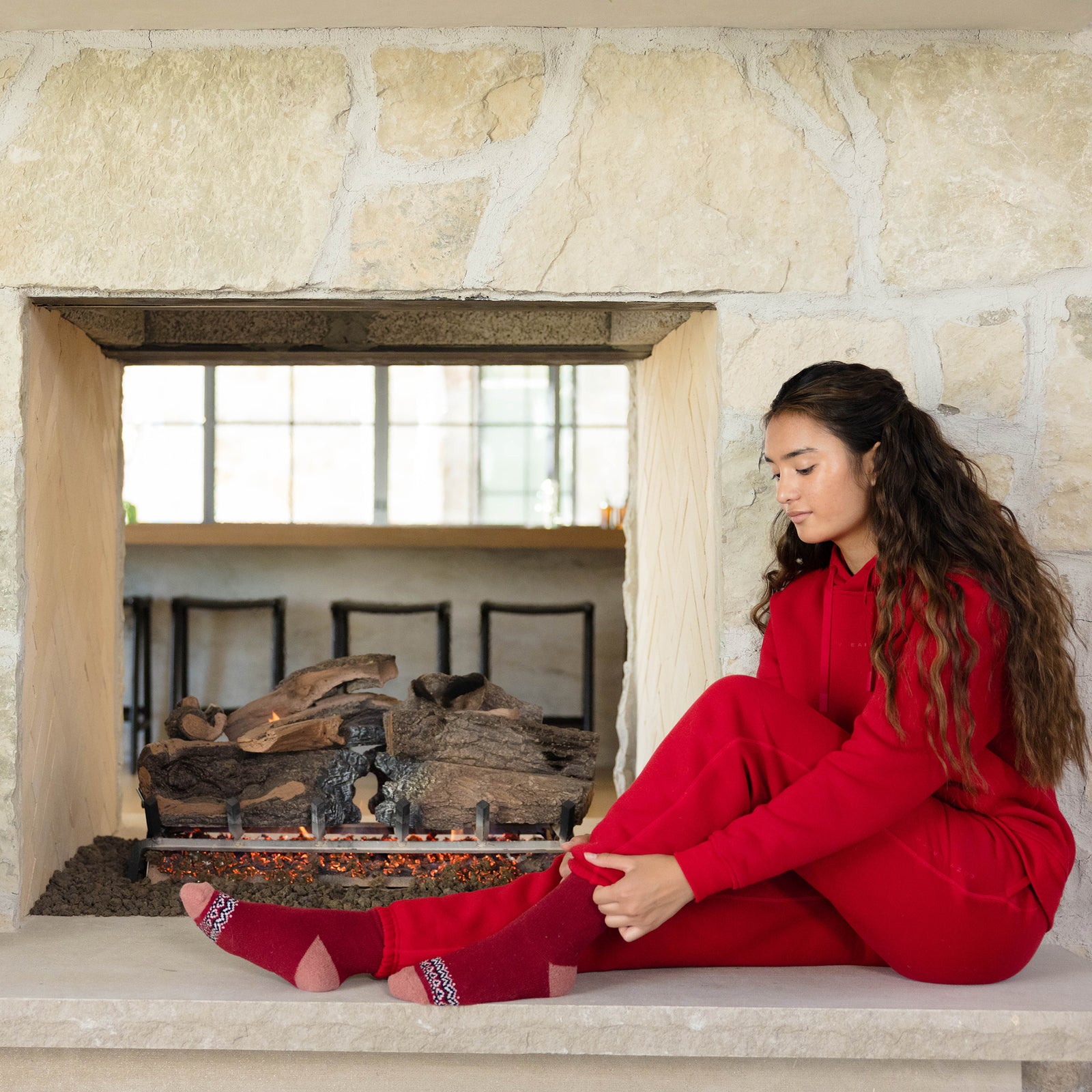 A child wearing a Kid's CityScape Hoodie by Cozy Earth sits on the stone hearth of an unlit fireplace, adjusting their red socks. The room has a cozy ambiance with a large window in the background.