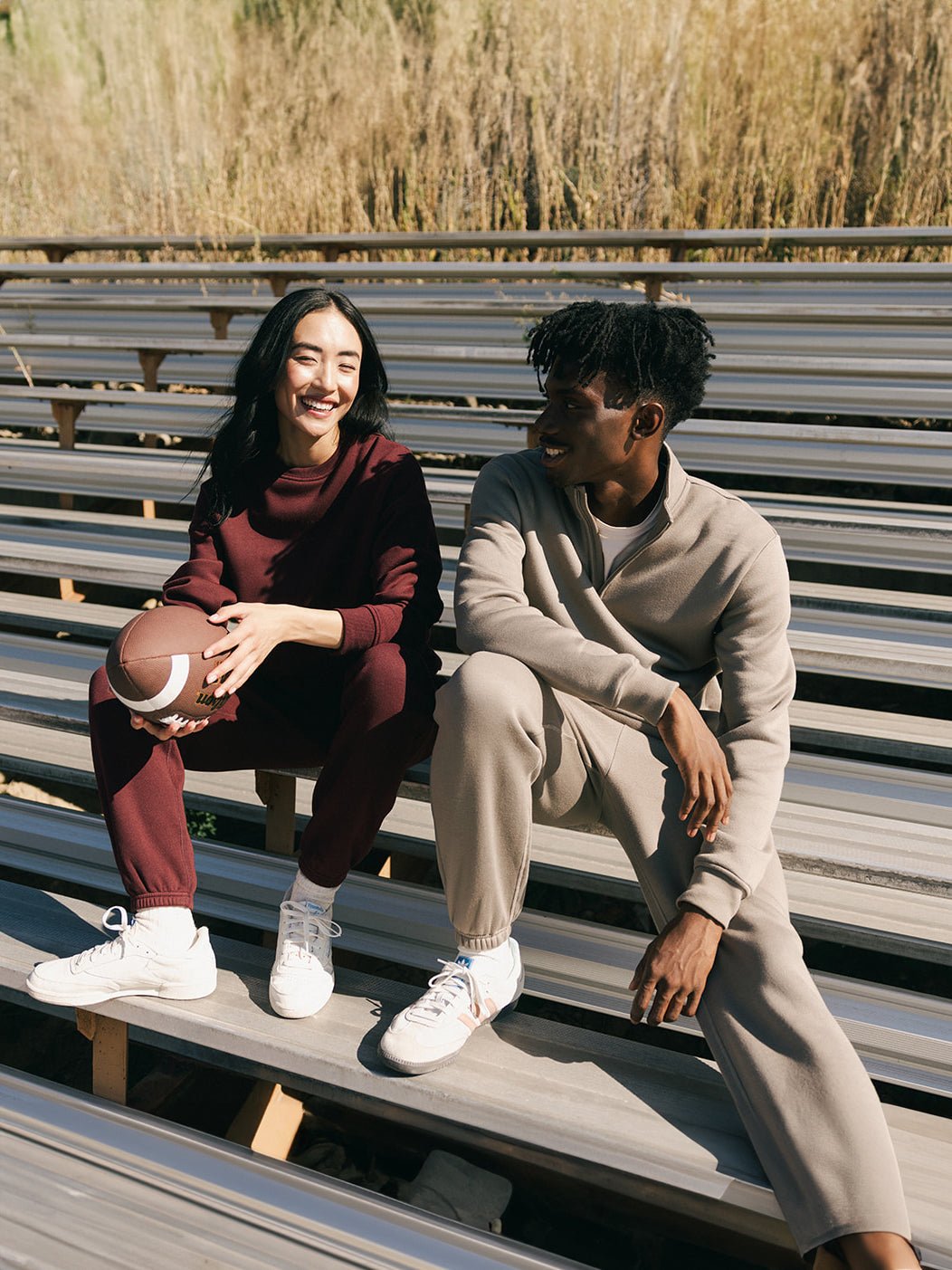 Two people sit on bleachers outside on a sunny day. The person on the left, wearing a red CityScape Quarter Zip by Cozy Earth, smiles while holding an American football. The person on the right, dressed in a beige CityScape Quarter Zip by Cozy Earth, smiles and looks at the other person. Both wear white sneakers.