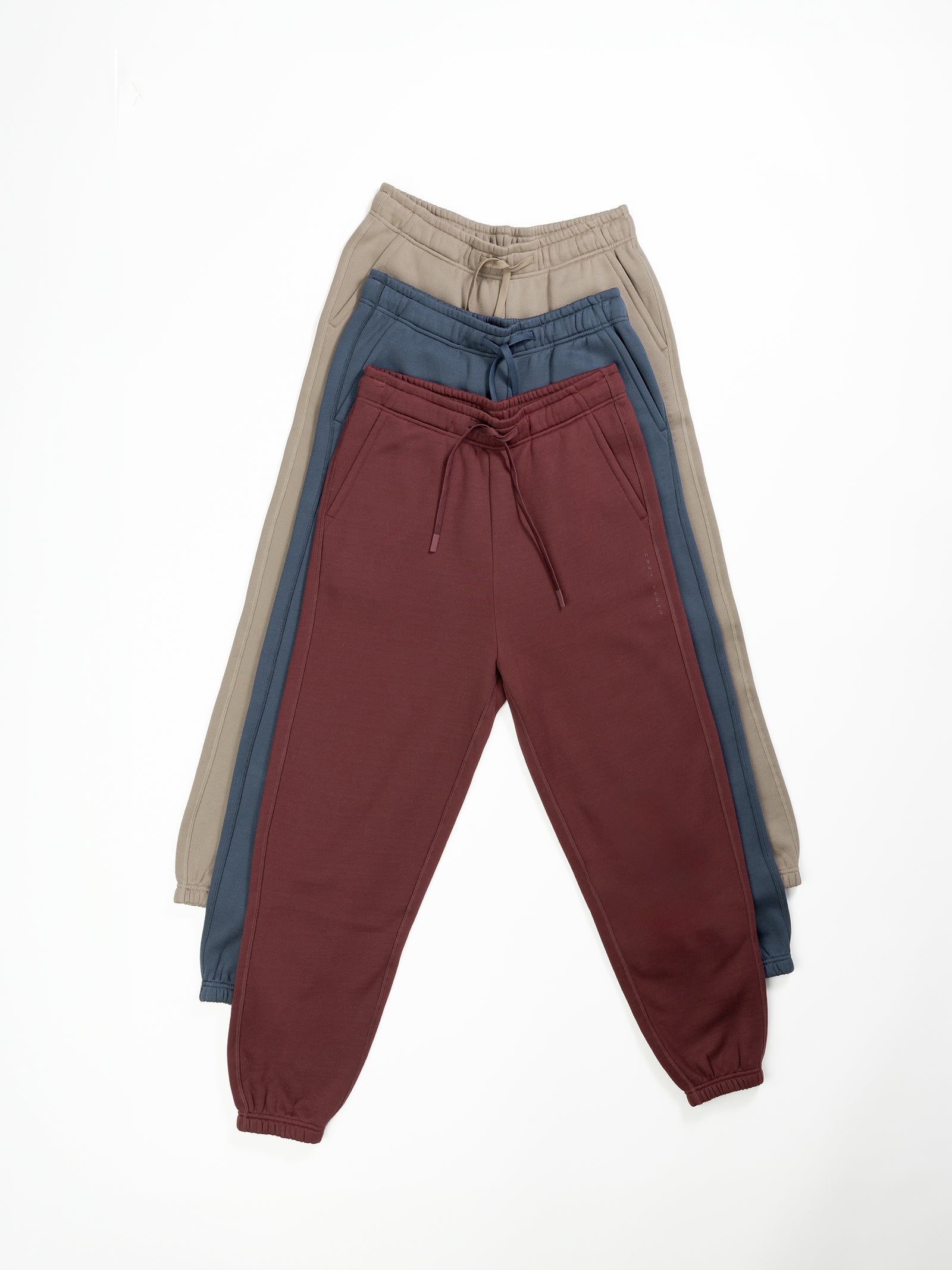 Three pairs of Cozy Earth's Men's CityScape Sweatpants are arranged in a fan layout on a white background. The front pair is maroon, the middle pair is blue, and the back pair is beige. All feature drawstrings and elastic cuffs.