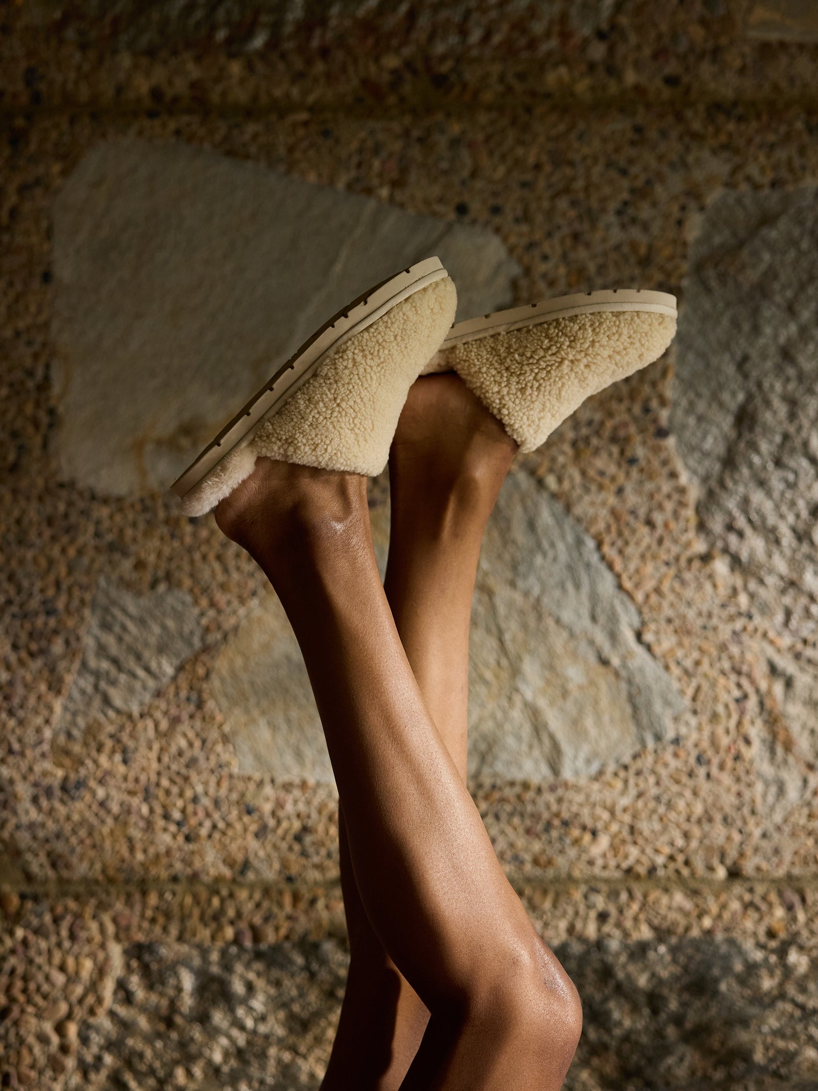 A person crosses their legs upwards, showcasing beige Puffy Sheep Slippers by Cozy Earth against a textured stone wall backdrop. The slippers have a cozy, soft appearance with sturdy soles. The ambient lighting casts gentle shadows, emphasizing the texture and warmth of the scene
