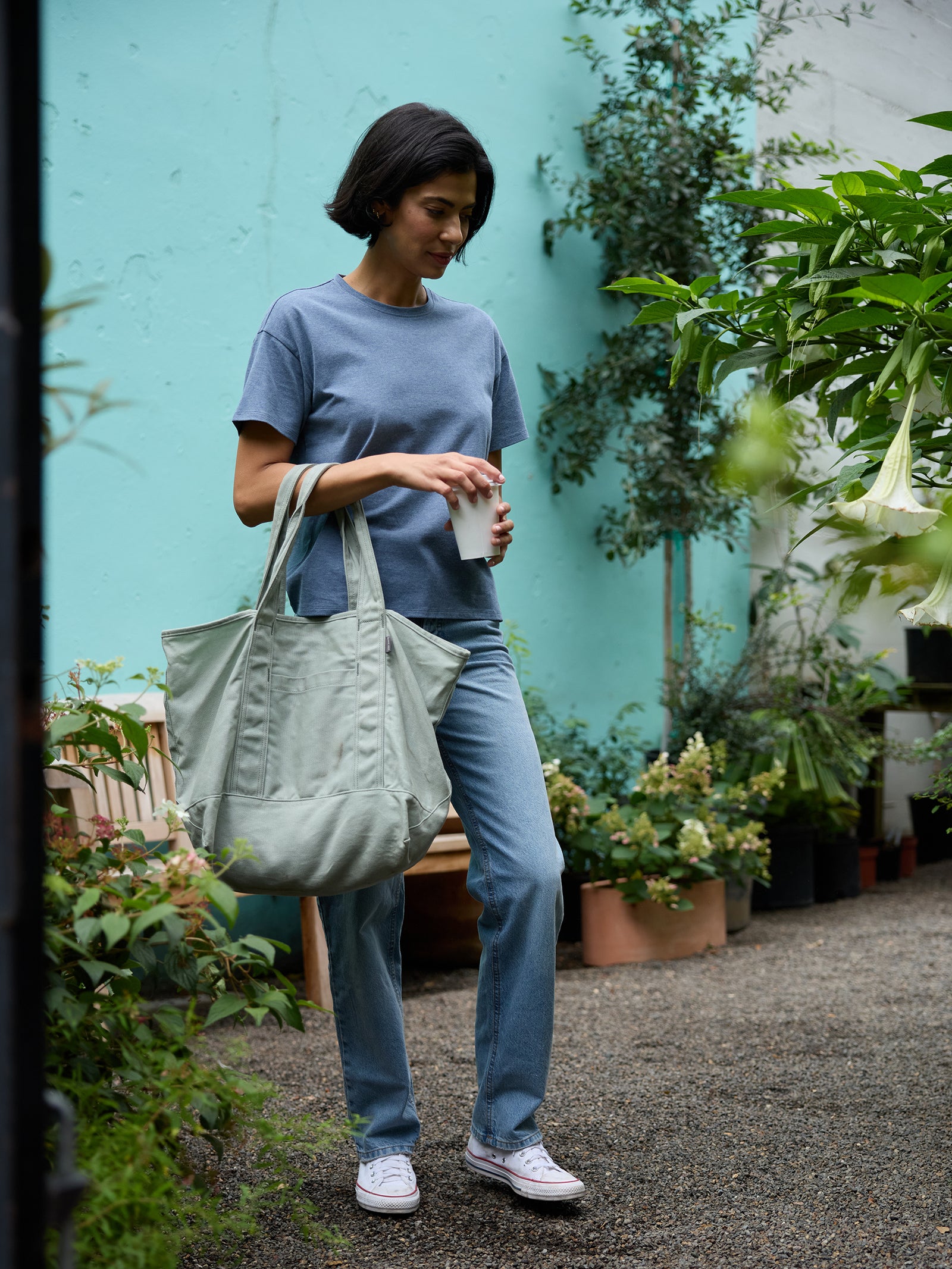 A person with short dark hair, wearing a blue shirt, jeans, and white sneakers, is standing outdoors holding a drink and a large green Waxed Canvas Tote from Cozy Earth. They are surrounded by plants and greenery, with a turquoise wall in the background.