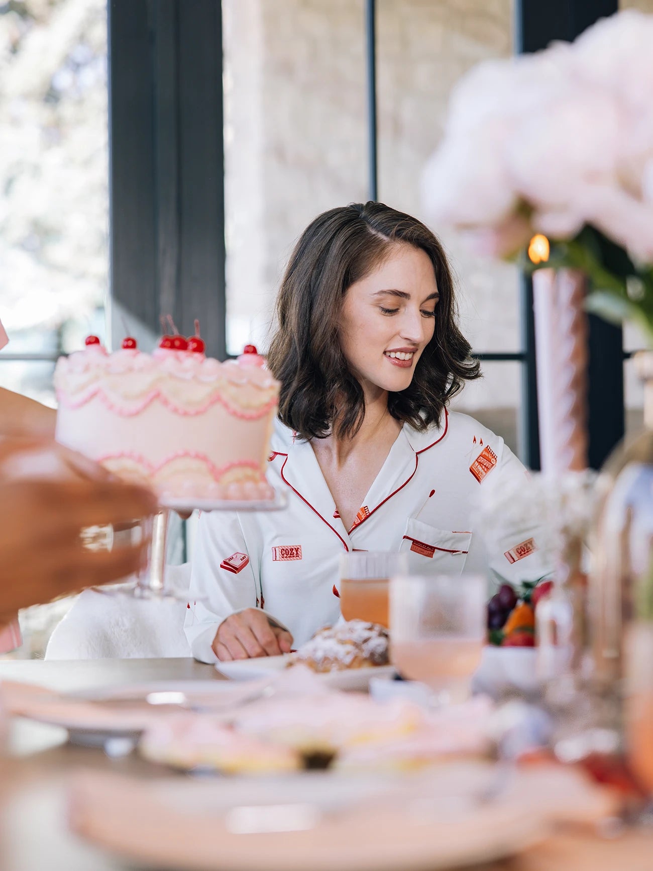 A woman wearing Cozy Earth Women's Long Sleeve Bamboo Pajama Top in Stretch-Knit smiles at a table with a cherry-topped pink frosted cake, surrounded by breakfast items and flowers. Bright daylight shines through the windows behind her.