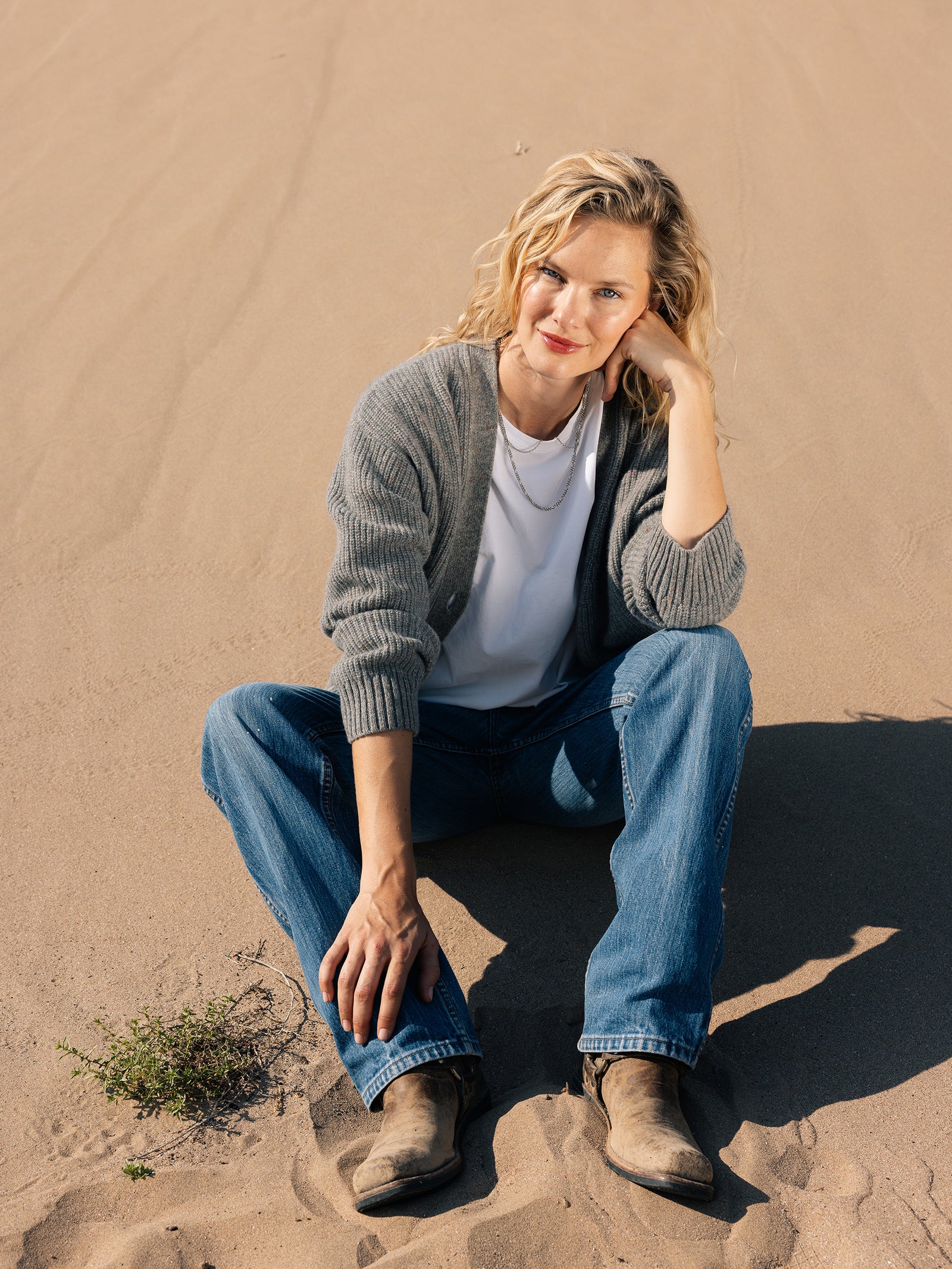 A person with curly blonde hair sits on a sandy ground, wearing an Oversized Sunday Cashmere Cardigan by Cozy Earth, along with a white shirt, blue jeans, and brown boots. They rest their head on their hand and smile at the camera. A small plant grows nearby.