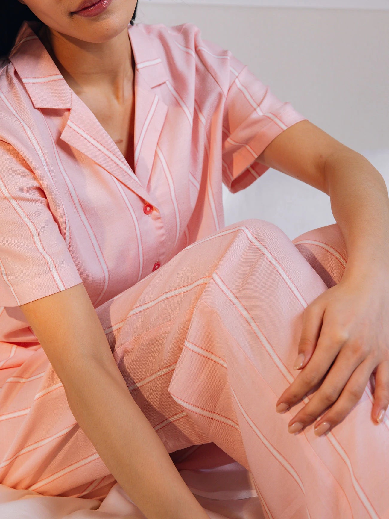 A person in pink striped pajamas sits on a bed with arms resting on their knees, showcasing the Women's Soft Woven Pajama Pant from Cozy Earth, featuring a relaxed fit and button-up shirt. The background is softly blurred.