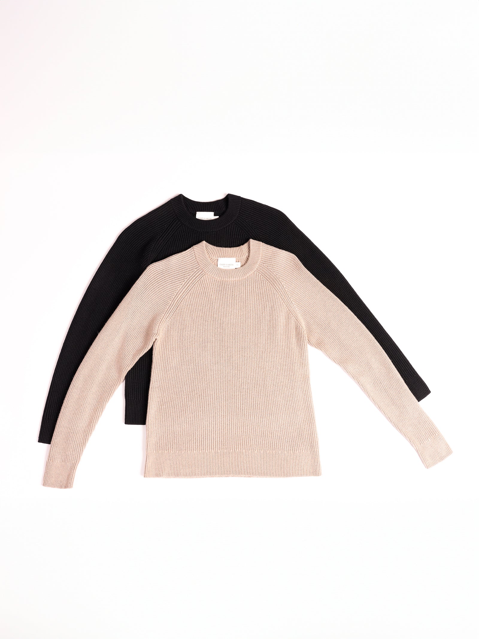 Two Women's Classic Crewneck sweaters from Cozy Earth are laid flat on a white surface. The front sweater is beige, and the back sweater is black. Both feature a crew neck design and a ribbed texture.