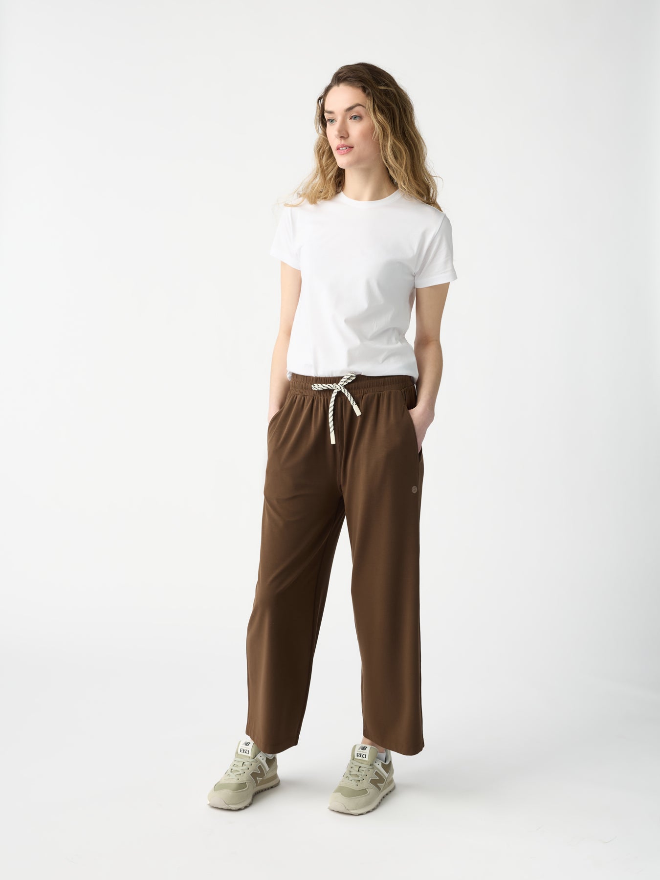 A woman in a white t-shirt and Cozy Earth's Women's Studio Cropped Wide Leg Pants, with hands in pockets, stands against a plain white background. She sports light sneakers and has long, wavy hair.