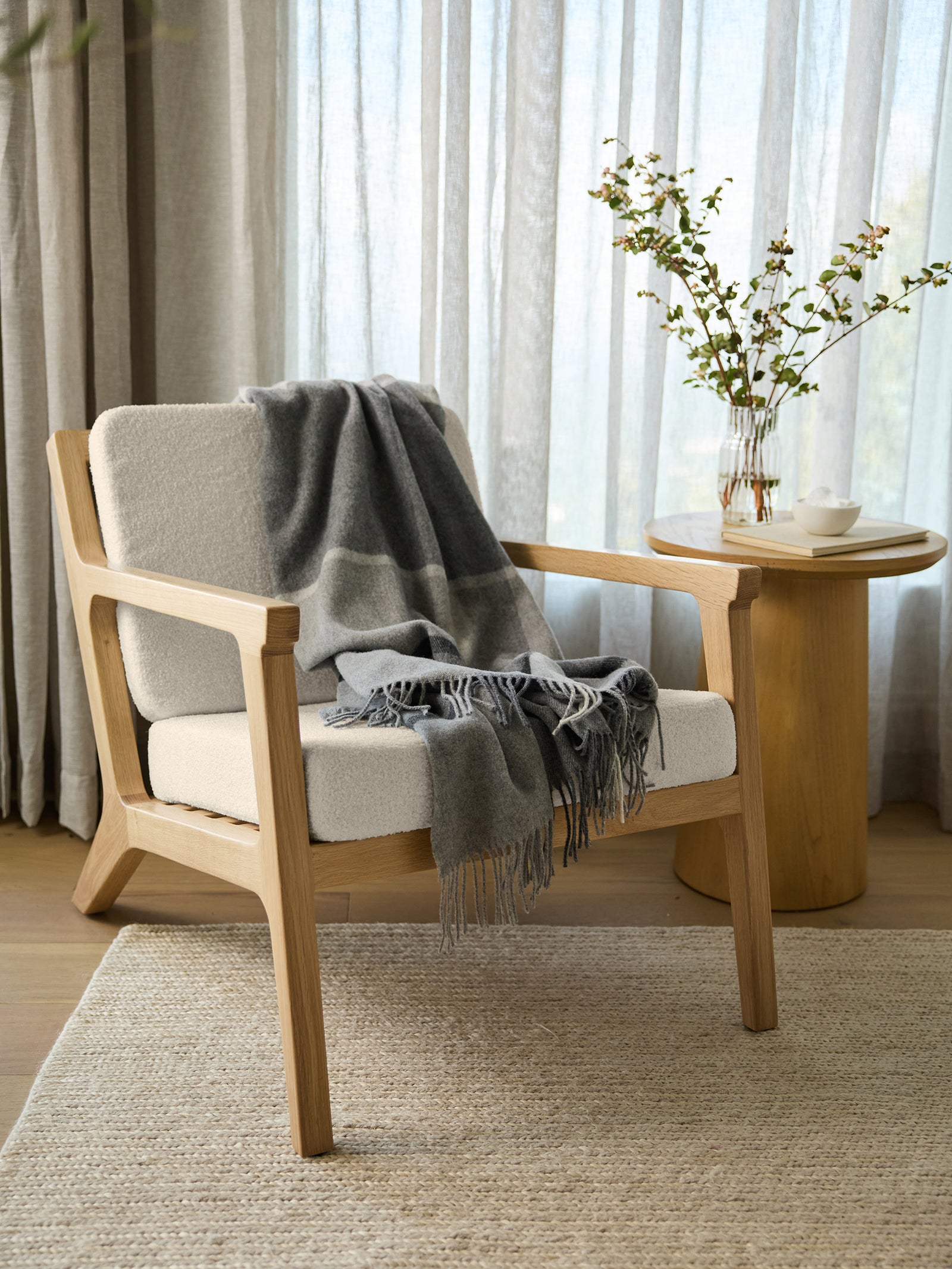 The Coronado Lounge Chair by Cozy Earth, adorned with a grey throw blanket, comfortably rests on a woven rug. Next to it stands a wooden side table that supports a vase filled with branches and white flowers, alongside a teacup. Sheer curtains softly diffuse natural light throughout the room.