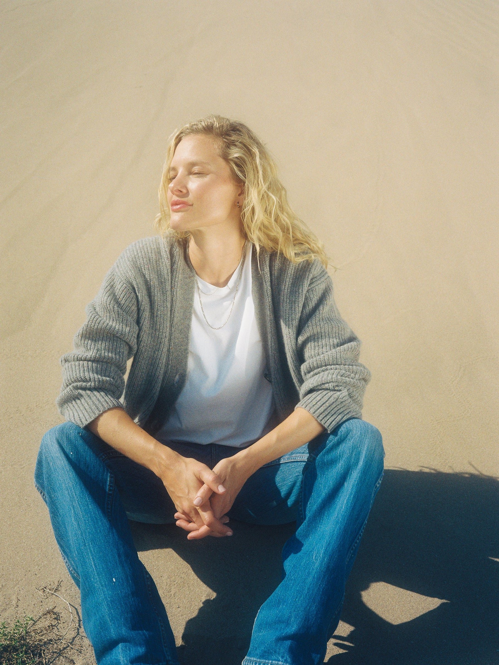 A person with long blonde hair sits cross-legged on the sand, wearing an Oversized Sunday Cashmere Cardigan by Cozy Earth, paired with a white shirt and blue jeans. They have their eyes closed and face tilted towards the sun, appearing relaxed and content.