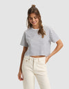 A woman with long brown hair wears the Cozy Earth Women's All Day Cropped Tee, featuring white and black stripes, and high-waisted cream jeans, standing against a light gray background with a smile and one hand in her pocket. |Color:Jet Black Stripe