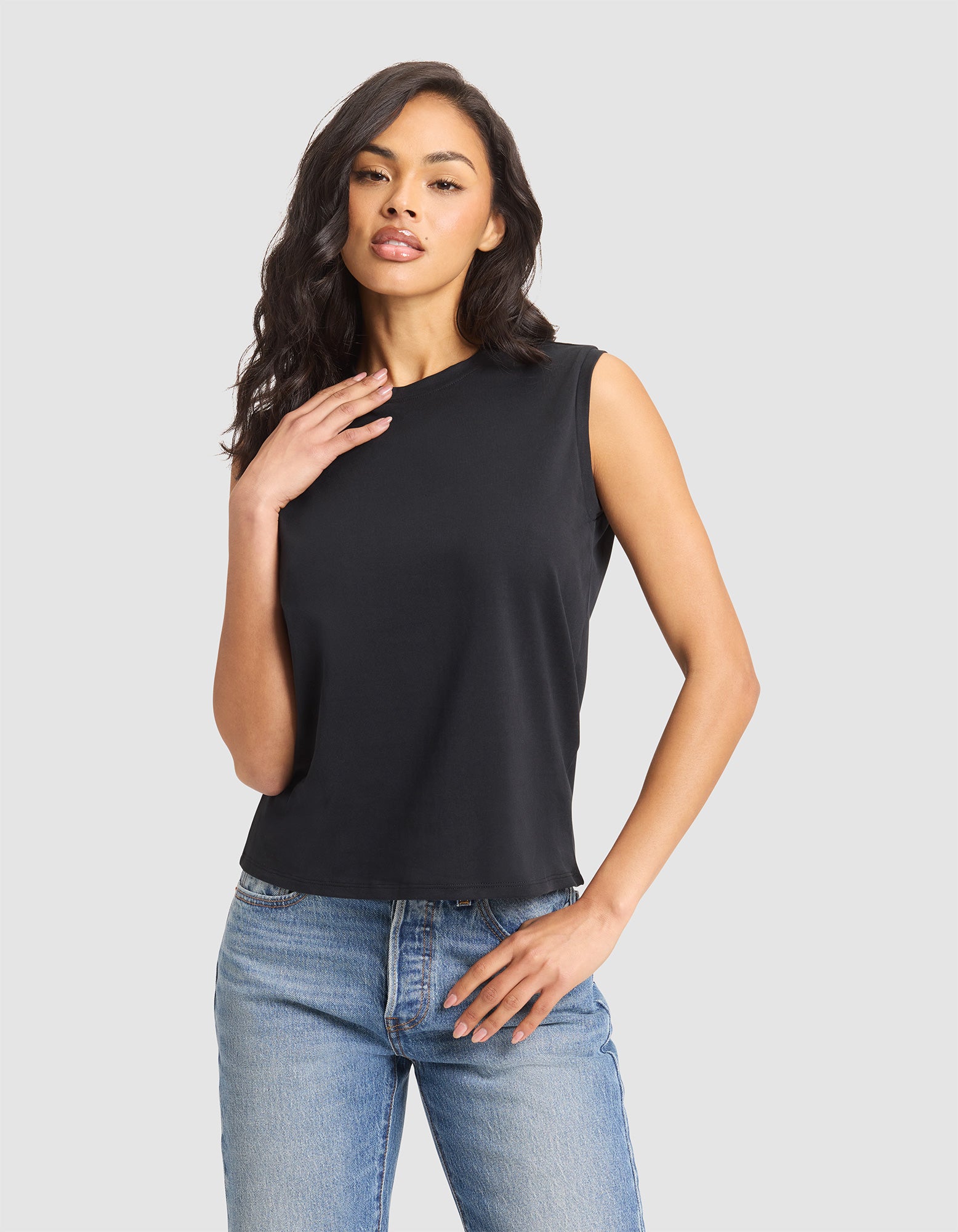 A woman models the Cozy Earth Women's All Day Sleeveless Tee in black, paired with light blue jeans, standing against a plain light gray background with one hand on her hip and the other touching her collarbone. |Color:Jet Black