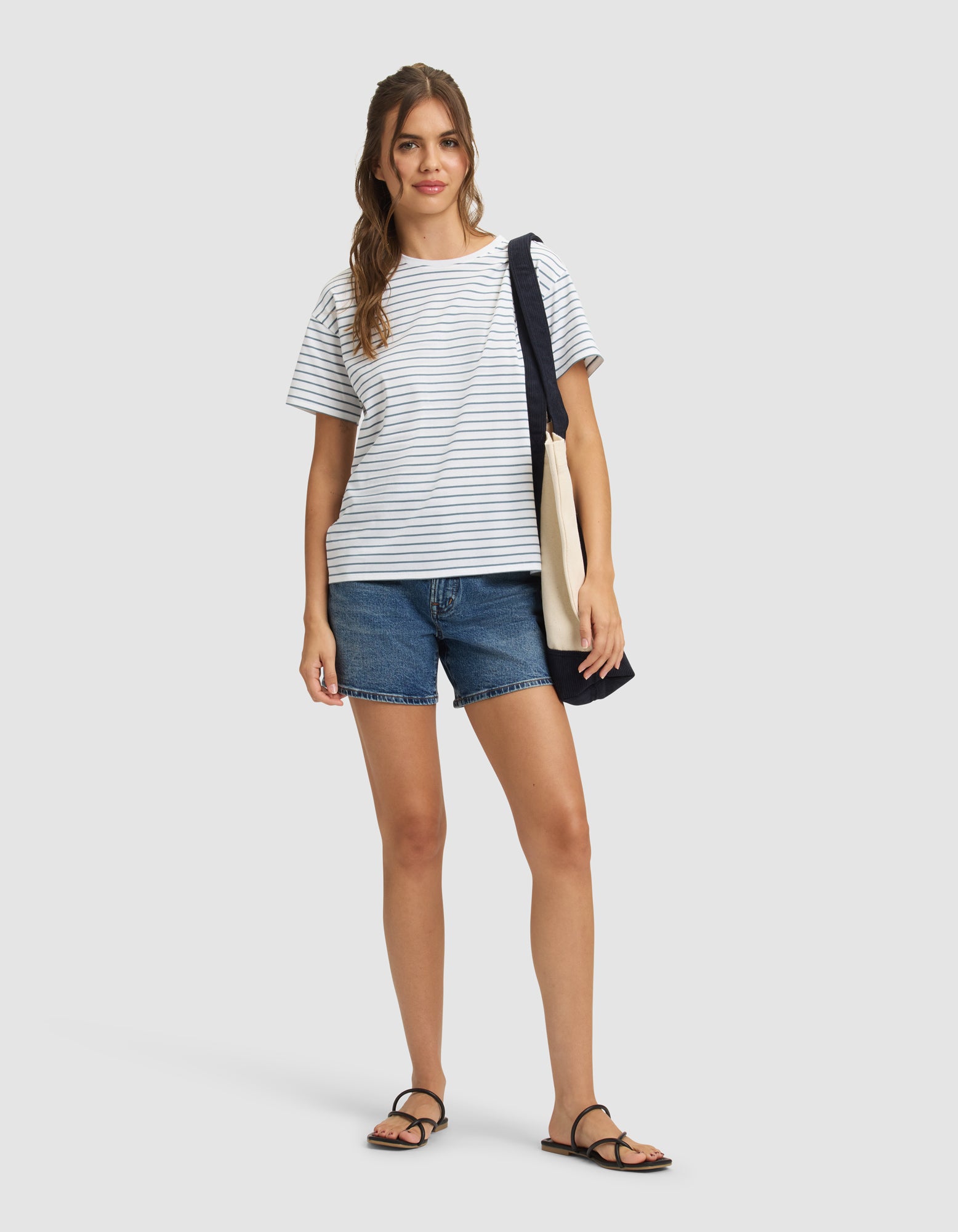 A woman with long, wavy hair stands against a plain background, smiling slightly and wearing the Cozy Earth Women's All Day Tee in white and gray stripes, paired with denim shorts, black sandals, and a navy-beige tote bag over her shoulder.