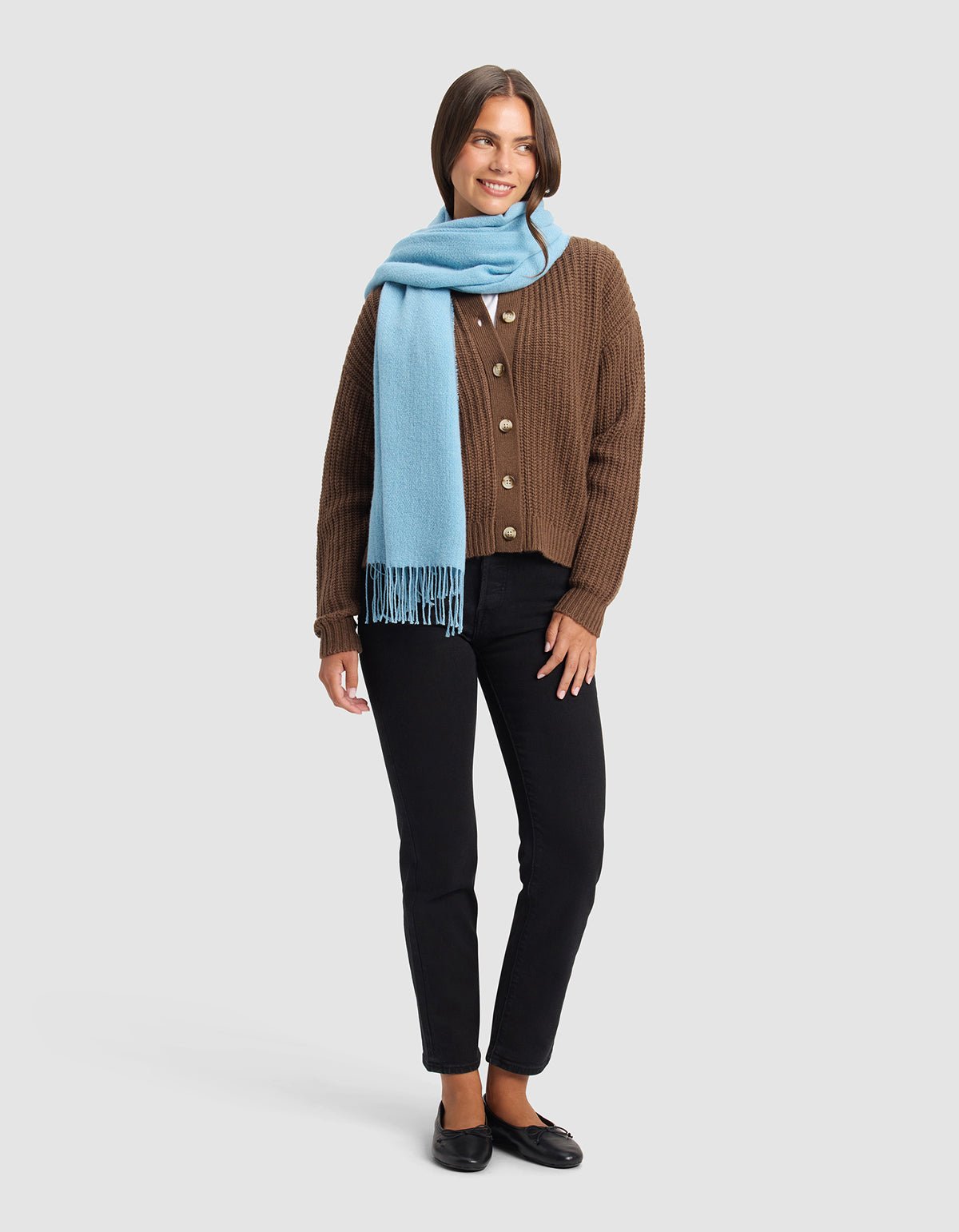 A smiling woman stands against a plain background, wearing the Cozy Earth Amelia Wrap in light blue around her neck, paired with a brown buttoned cardigan, black pants, and black shoes.