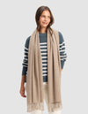 A woman stands against a light gray background wearing the Cozy Earth Amelia Wrap, a long beige scarf with fringed ends, paired with a blue and white striped sweater and cream-colored pants. |Color:Taupe