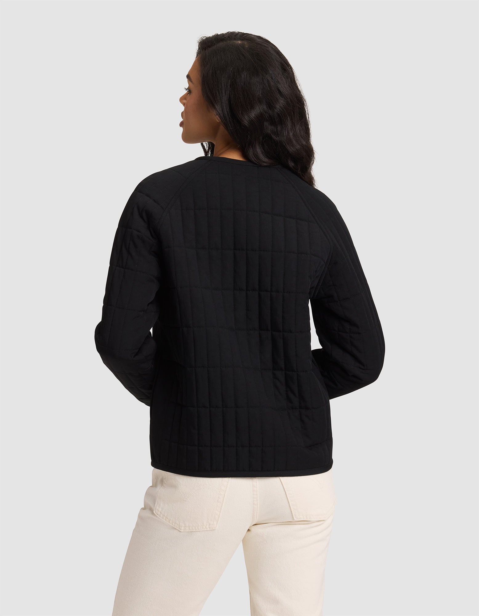 A woman is seen from the back wearing the Cozy Earth Women's Andie Quilted Jacket in black with light pants, standing against a plain light gray background.