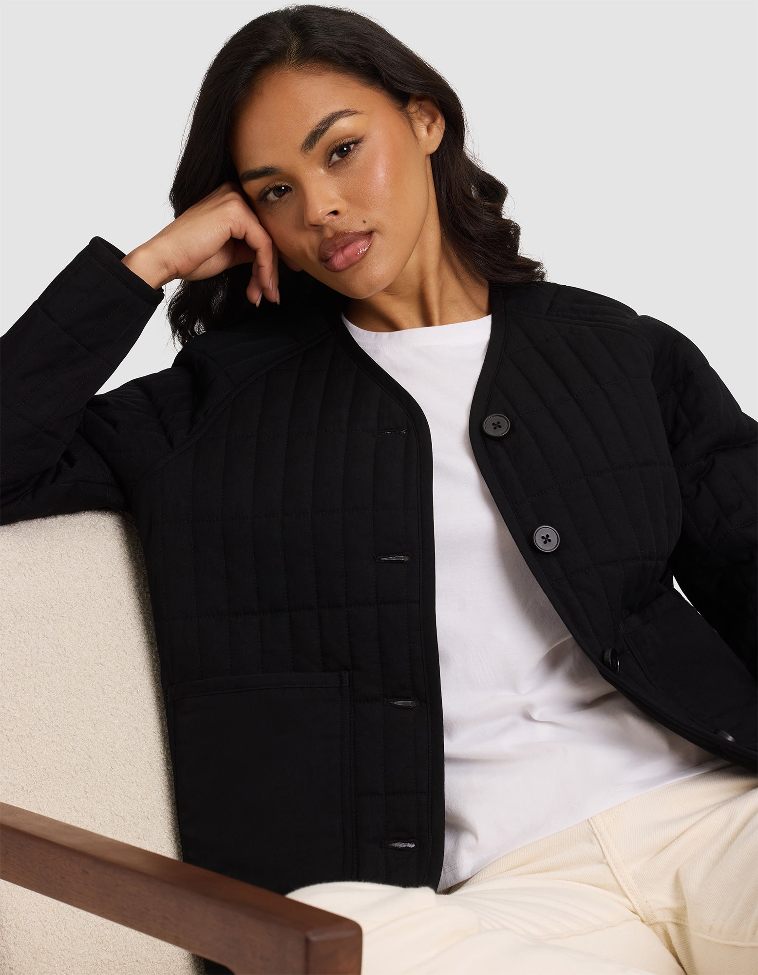 A woman with long dark hair sits on a chair, calmly looking at the camera. She wears the Cozy Earth Women's Andie Quilted Jacket over a white top and light pants, set against a plain background.