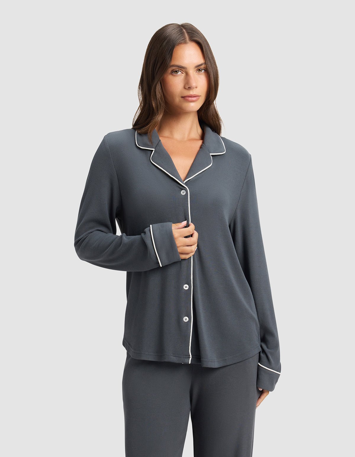 A woman with long brown hair wears the Cozy Earth Women's Bamboo Rib-Knit Classic Long Sleeve Pajama Top in dark gray with white piping and buttons, standing against a plain light gray background, holding one side of the top.|Color:Graphite