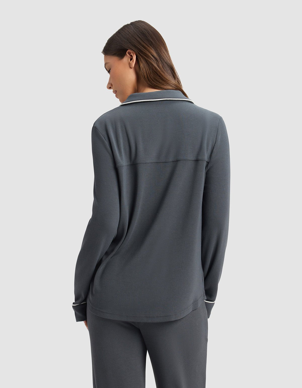 A woman with long brown hair stands facing away, wearing the Cozy Earth Women's Bamboo Rib-Knit Classic Long Sleeve Pajama Top in dark gray against a light gray background.