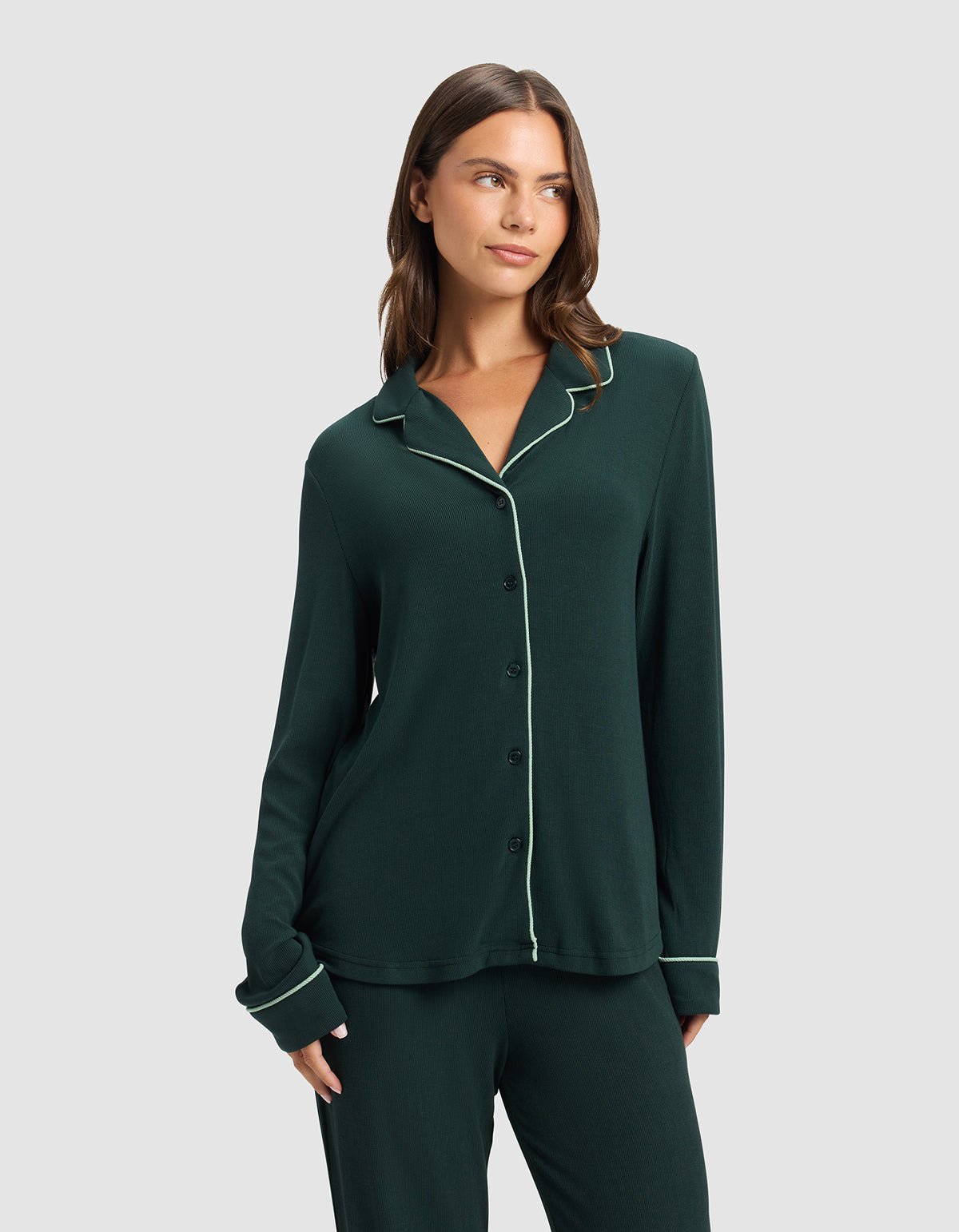 A woman wears the Cozy Earth Women's Bamboo Rib-Knit Classic Long Sleeve Pajama Top, dark green with white piping, standing against a plain light gray background and looking to the side with one hand in her pocket.|Color:Spruce