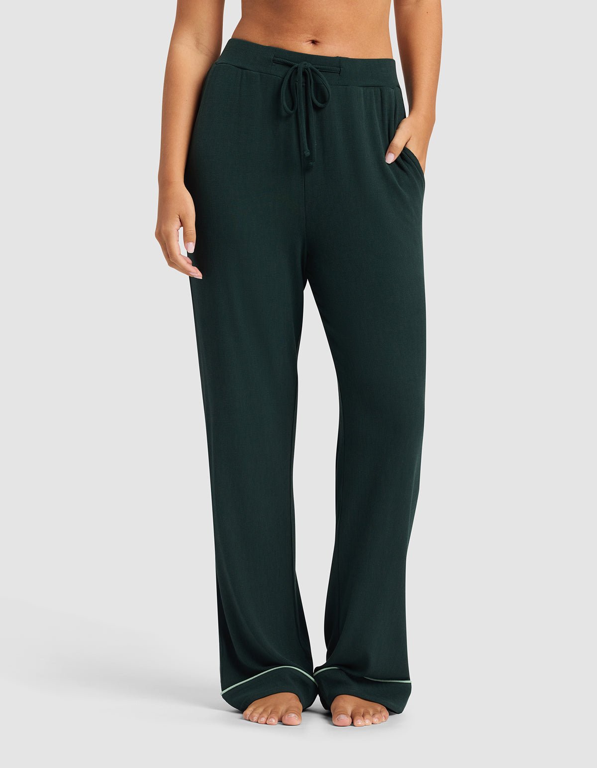 A woman wears Cozy Earth's Women's Bamboo Rib-Knit Classic Pajama Pant in dark green, stands barefoot with hands in pockets against a plain light gray background.|Color:Spruce