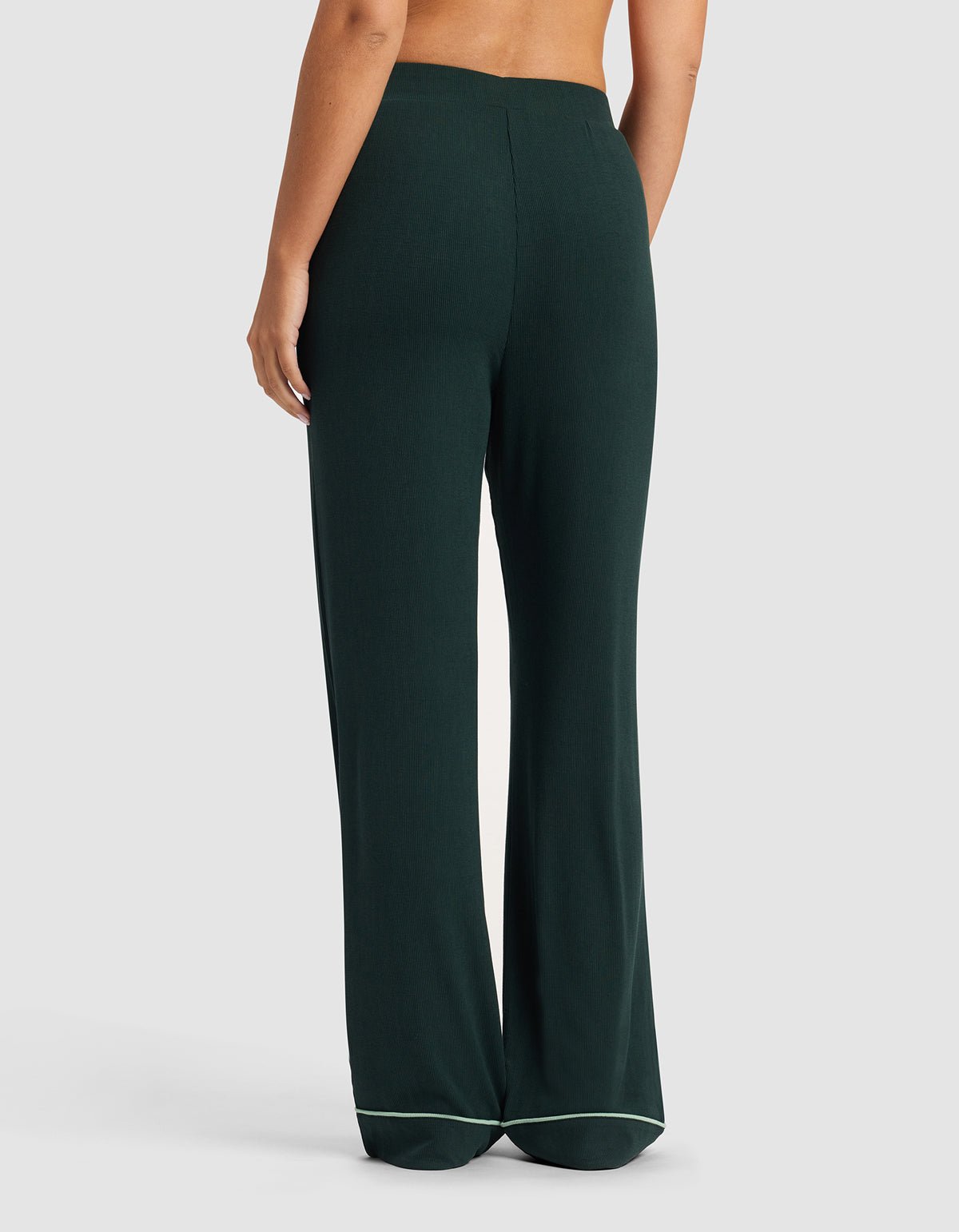 A person stands against a plain background, photographed from the back, wearing Cozy Earth's Women's Bamboo Rib-Knit Classic Pajama Pant in dark green with a light-trimmed hem, showcasing the full length and wide-leg, high-waisted fit.