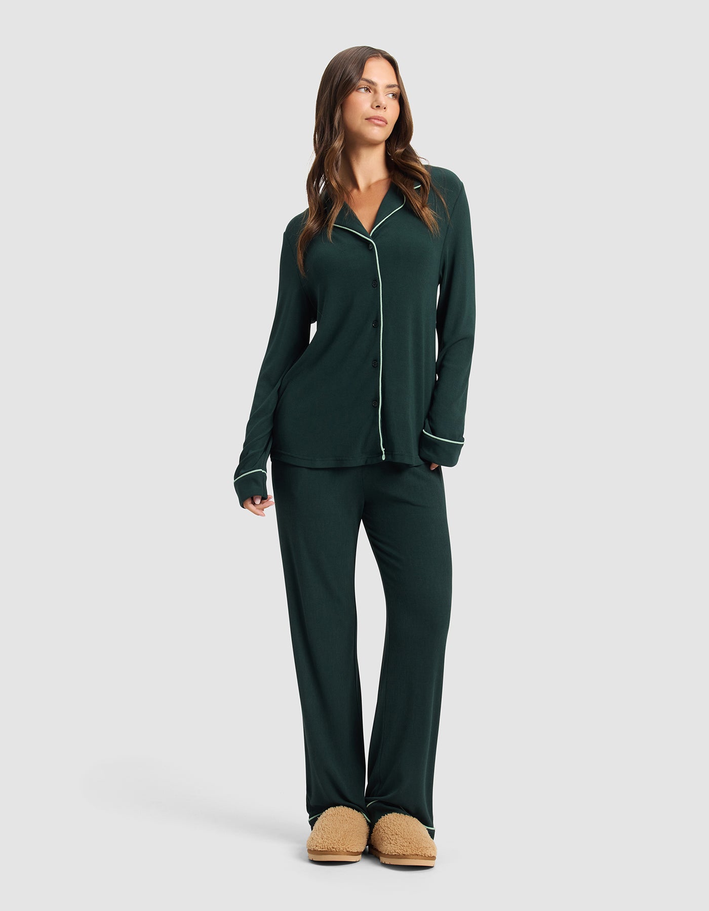 A woman in the Cozy Earth Women's Bamboo Rib-Knit Classic Long Sleeve Pajama Set in dark green with white piping and tan slippers stands against a light gray background, gazing slightly aside with a relaxed expression.|Color:Spruce