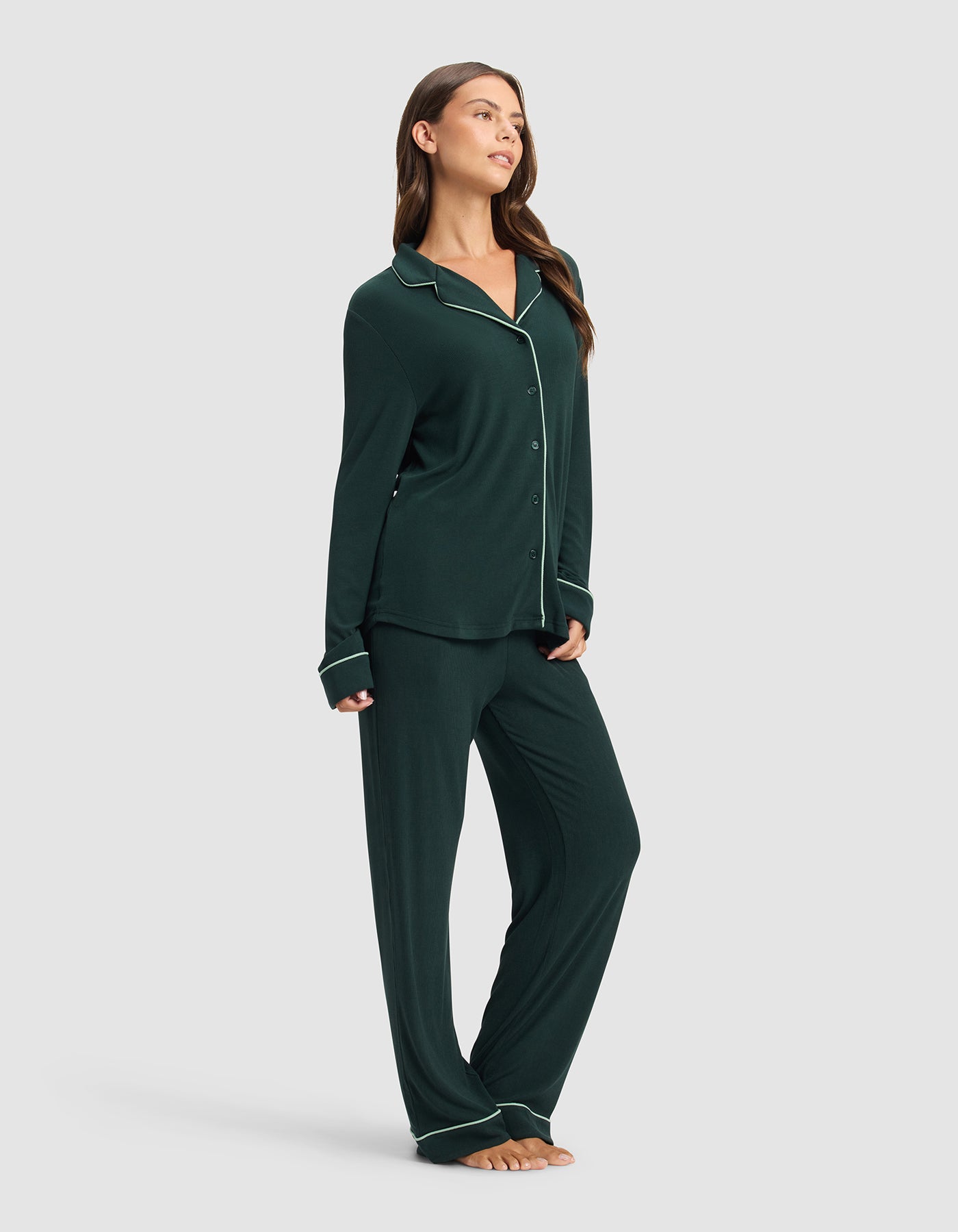 A woman stands barefoot, smiling slightly and looking to the side, wearing Cozy Earth's Women's Bamboo Rib-Knit Classic Long Sleeve Pajama Set in dark green with white piping, against a plain light gray background.