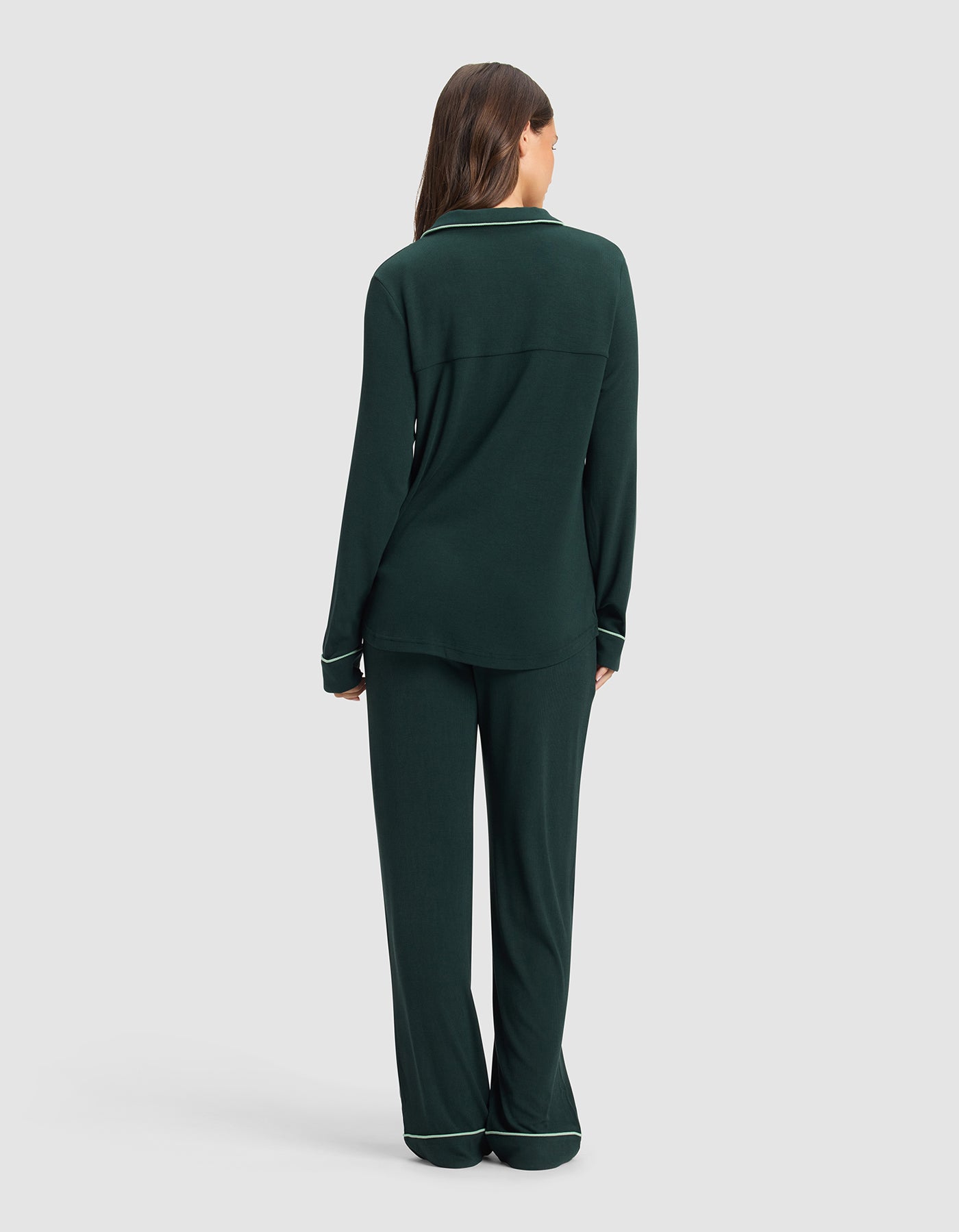 A woman with long brown hair, seen from behind, wears the Cozy Earth Women's Bamboo Rib-Knit Classic Long Sleeve Pajama Set in dark green with light piping, standing against a plain light gray background.