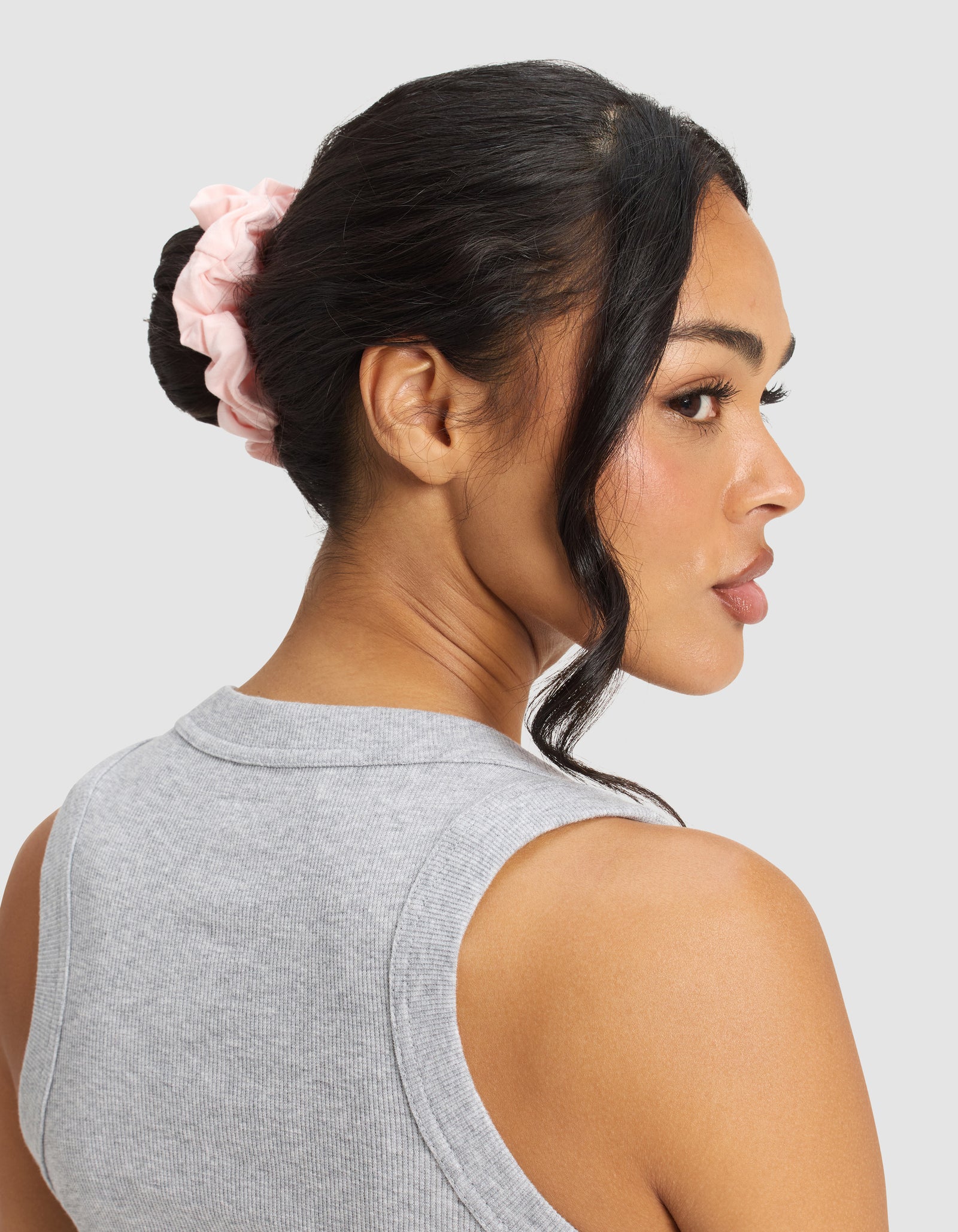 A woman with dark hair in a bun secured by a Cozy Earth Bamboo Stretch-Knit Scrunchie in light pink, wearing a sleeveless gray top, looks over her shoulder against a light gray background.
