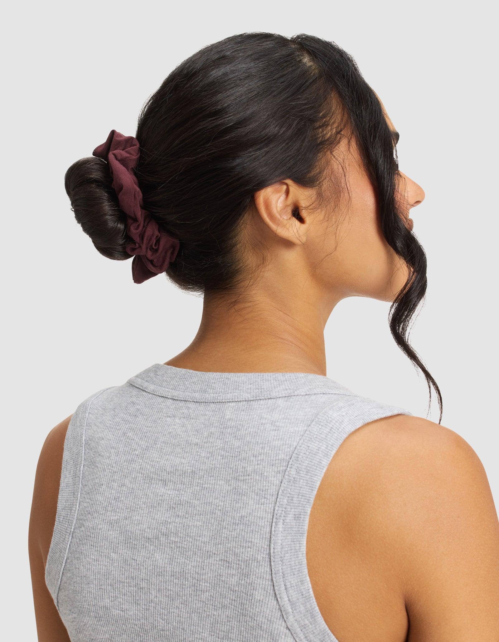 A woman with dark hair in a low bun secured by a Cozy Earth Bamboo Stretch-Knit Scrunchie is shown from behind, wearing a light gray sleeveless ribbed top against a plain background.