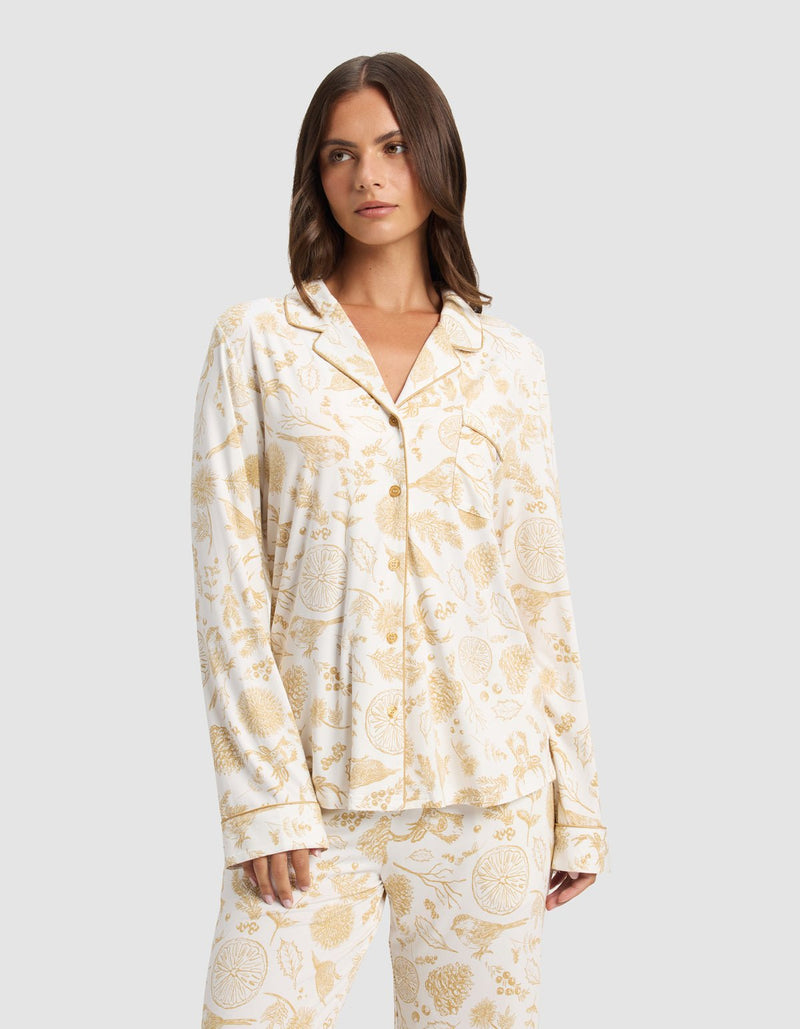 A woman wears the Cozy Earth Women's Bamboo Stretch-Knit Long Sleeve Pajama Top with a white base and light brown botanical and animal print, standing against a plain light gray background.