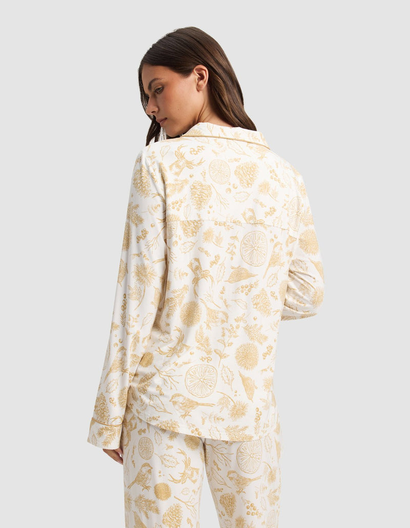 A woman with long brown hair is seen from behind wearing the Cozy Earth Women's Bamboo Stretch-Knit Long Sleeve Pajama Top in white with a yellow botanical and fruit print, standing against a light gray background.