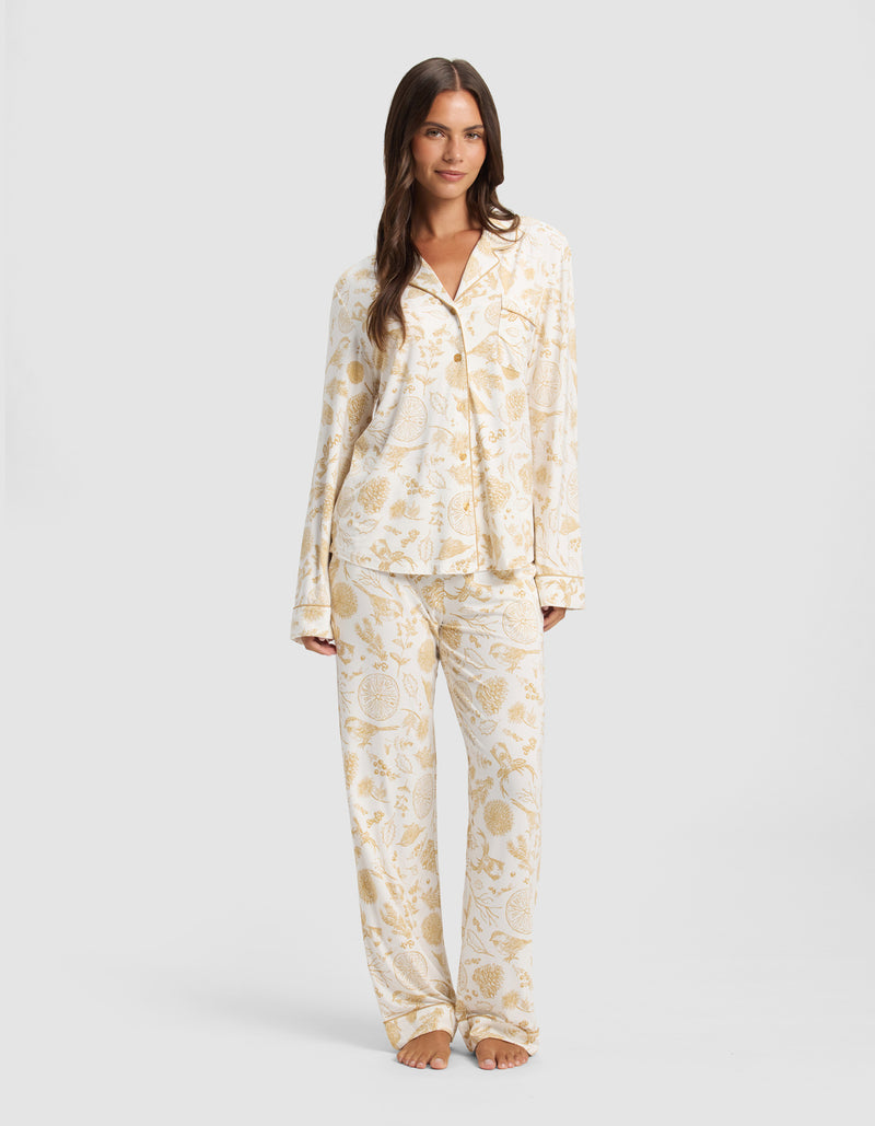 A woman stands barefoot against a plain light background, wearing Cozy Earth's Women's Bamboo Stretch-Knit Long Sleeve Pajama Top in cream with a gold floral and paisley pattern, paired with matching full-length pants.