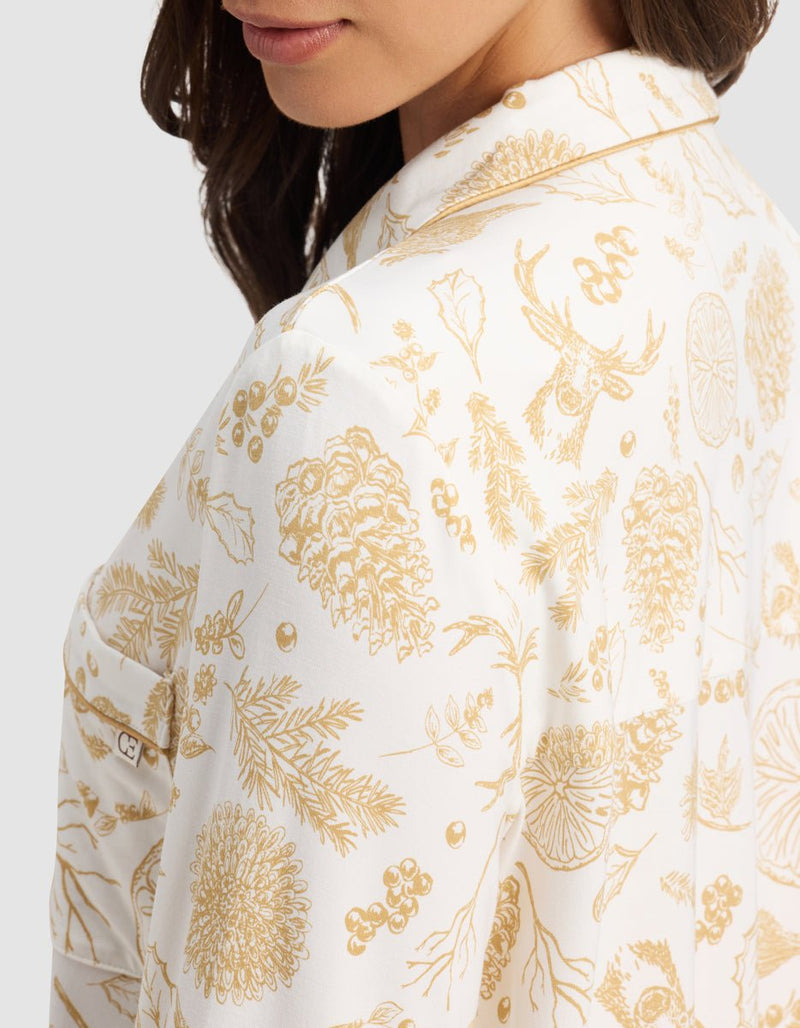 A woman wears the Cozy Earth Women's Bamboo Stretch-Knit Long Sleeve Pajama Top, featuring a white base with gold botanical and woodland animal prints—pinecones, sprigs, berries, and a stag's head. Side view highlights the shirt's detailed pattern.
