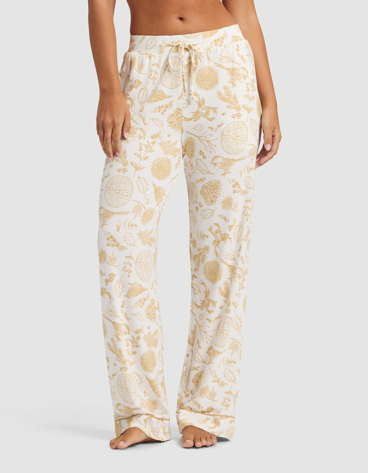 A person stands barefoot against a plain light background wearing Cozy Earth's Women's Bamboo Stretch-Knit Pajama Pant in a white color with a yellow floral and fruit pattern.