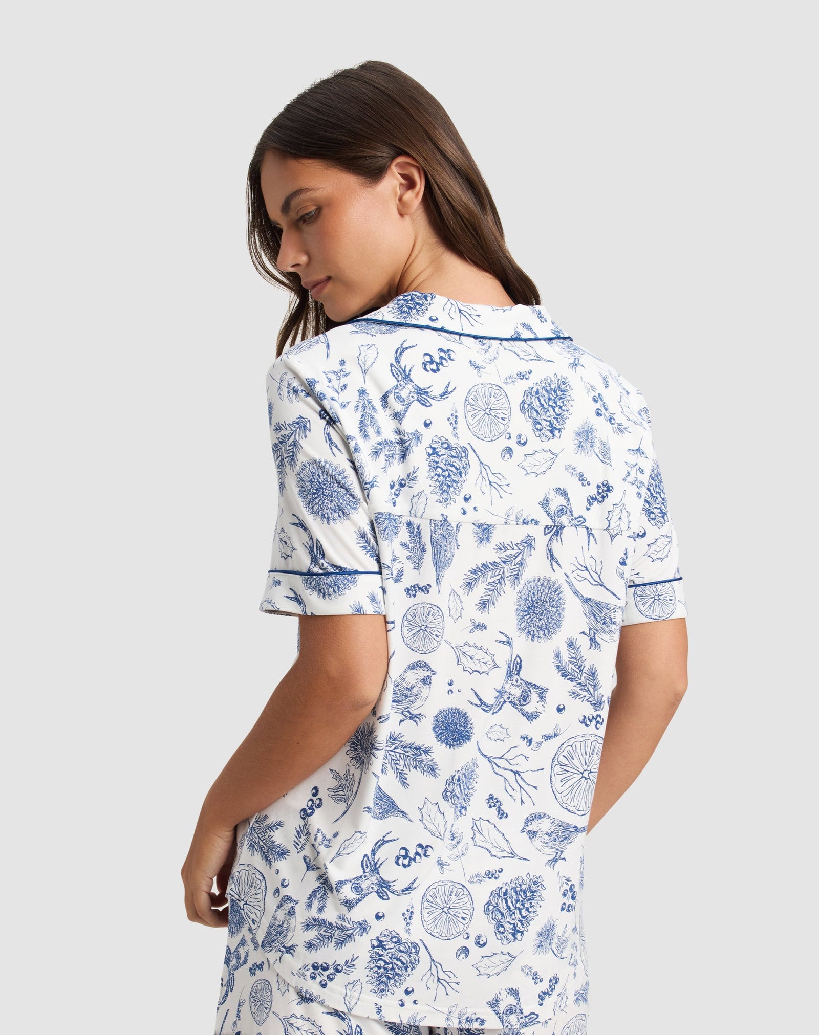 A woman stands turned slightly away, wearing the Cozy Earth Women's Bamboo Stretch-Knit Short Sleeve Pajama Top in white with blue botanical prints, a collar, and matching shorts. The background is plain light gray.