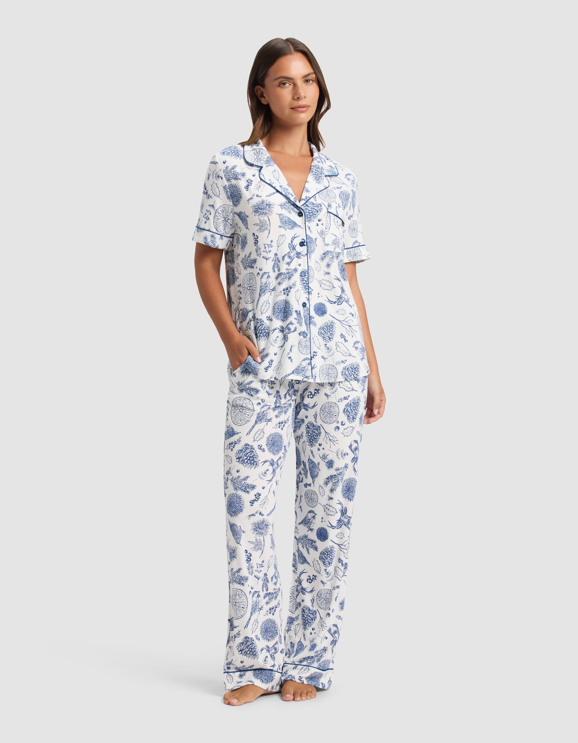 A woman stands barefoot in the Cozy Earth Women's Bamboo Stretch-Knit Short Sleeve Pajama Set, featuring a blue and white botanical print with a collared button-up top and loose pants. She has one hand in her pocket and looks relaxed.