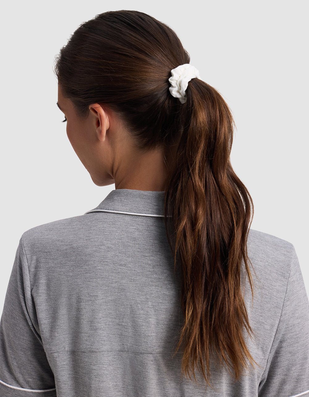 A woman with long brown hair in a ponytail secured by a white Cozy Earth Bamboo Stretch Knit Skinny Scrunchie, wearing a grey collared shirt, is shown from behind against a plain light background.