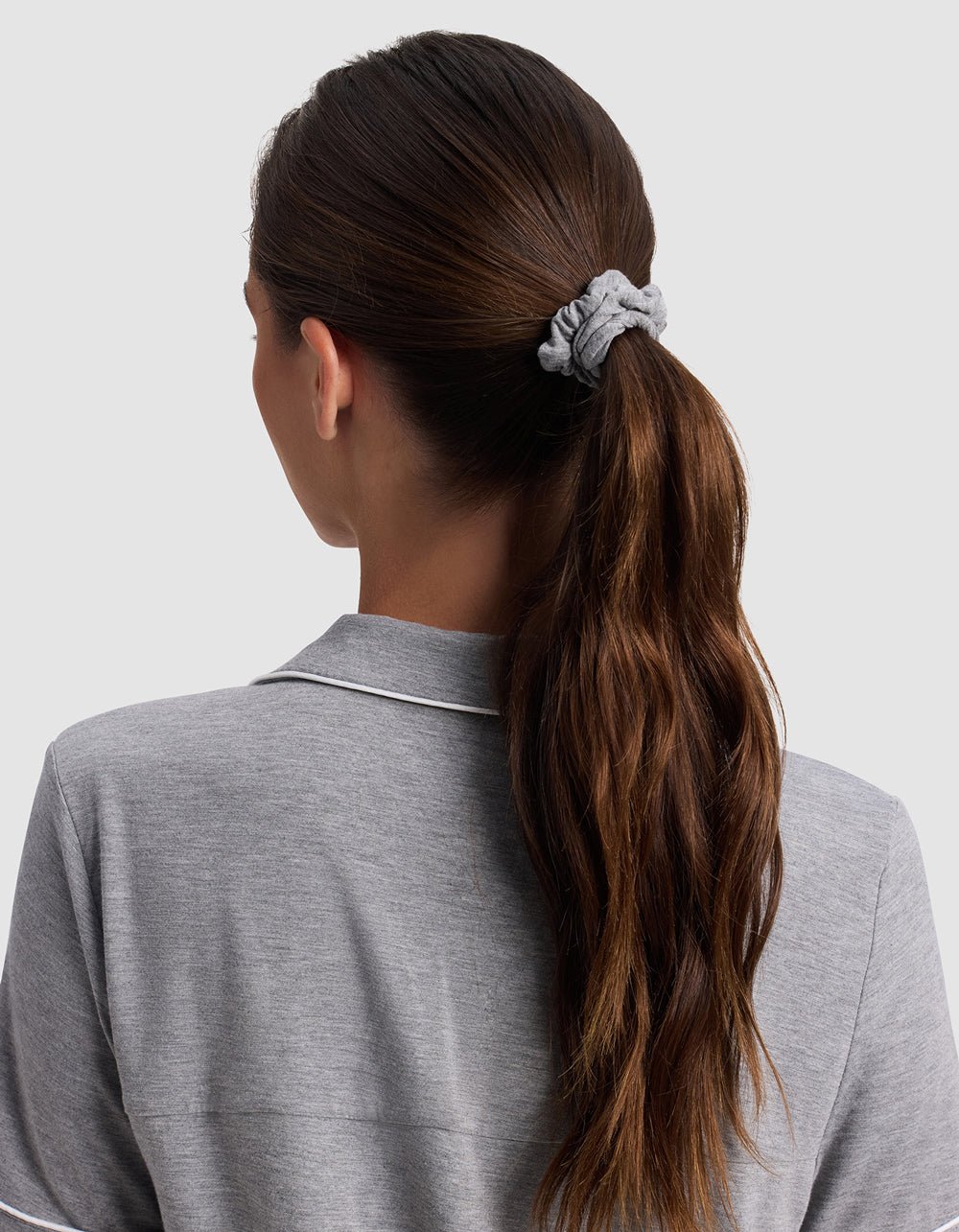 A woman with long brown hair in a ponytail, secured with a Cozy Earth Bamboo Stretch Knit Skinny Scrunchie, is shown from the back wearing a light gray collared shirt with white piping against a plain light background.