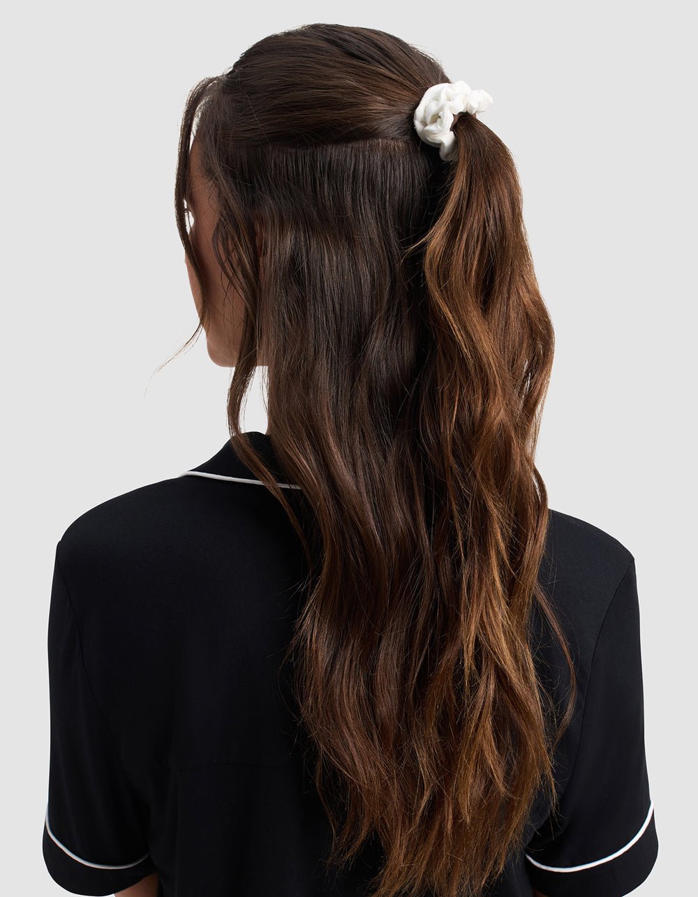 A woman with long, wavy brown hair in a half-up ponytail secured by a Cozy Earth Bamboo Stretch Knit Skinny Scrunchie is seen from the back, wearing a black top with white piping against a light background.