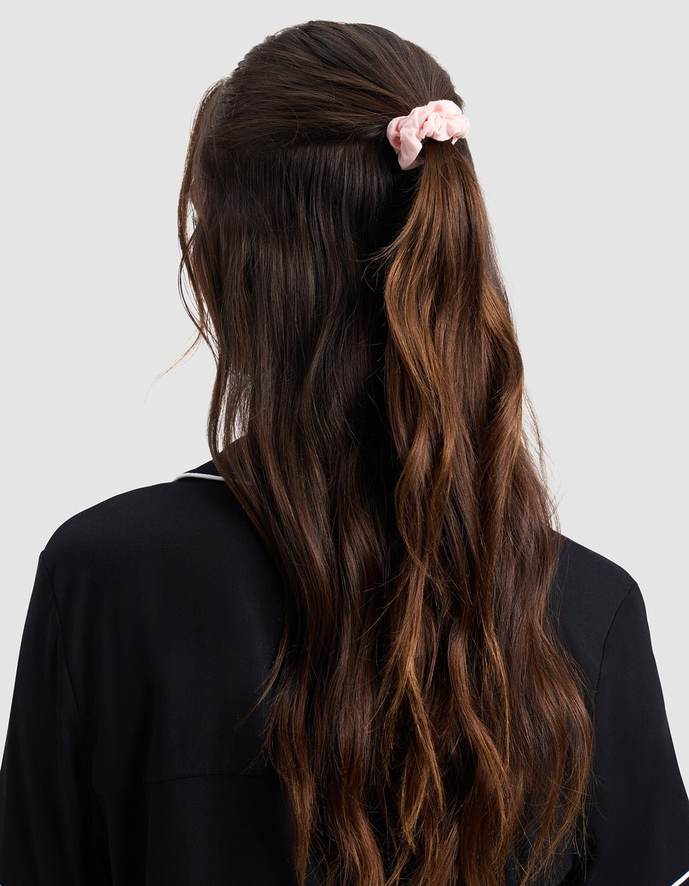 A woman with long, wavy brown hair wears a black top and secures part of her hair with a light pink Bamboo Stretch Knit Skinny Scrunchie by Cozy Earth, standing against a plain light gray background.