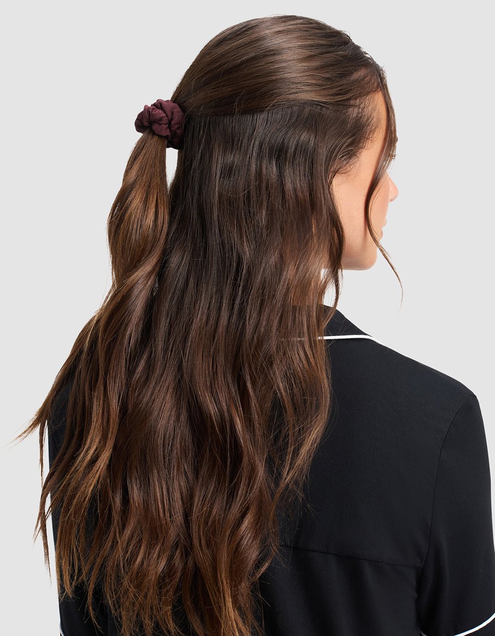 A woman with long, wavy brown hair is shown from behind, wearing a black top with white piping and her hair partially pulled back with a Cozy Earth Bamboo Stretch Knit Skinny Scrunchie.