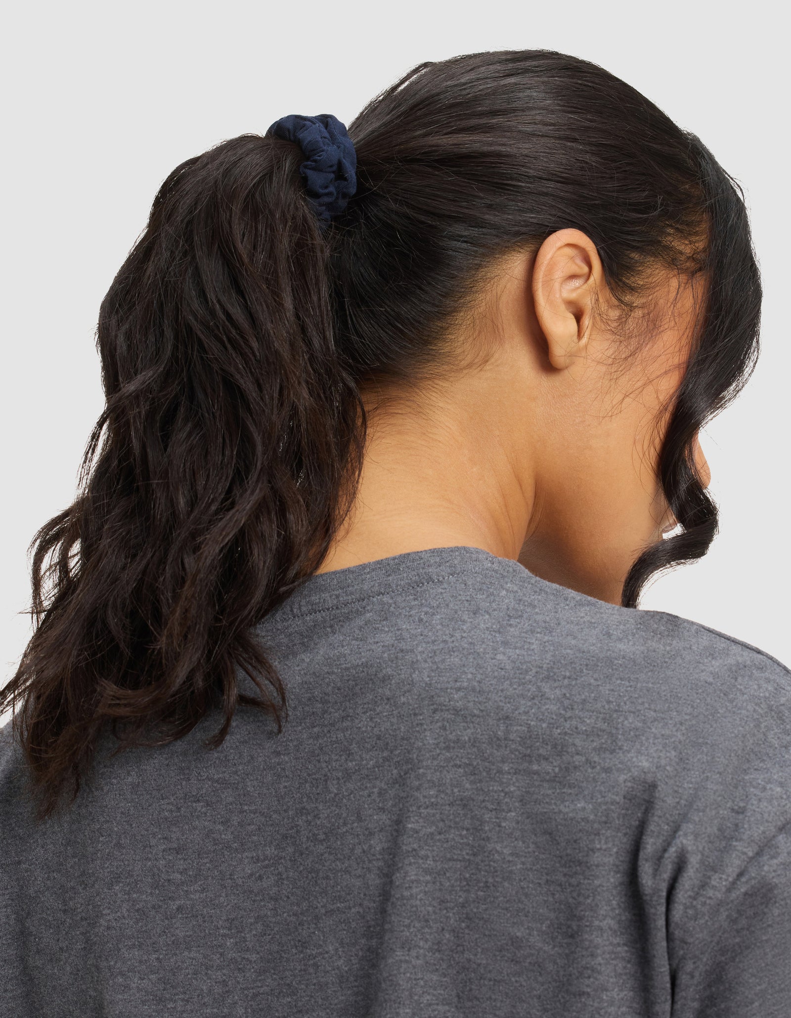 A person with long, dark, wavy hair tied in a ponytail with a Cozy Earth Bamboo Stretch Knit Skinny Scrunchie in navy, wearing a gray shirt, shown from behind against a plain background.