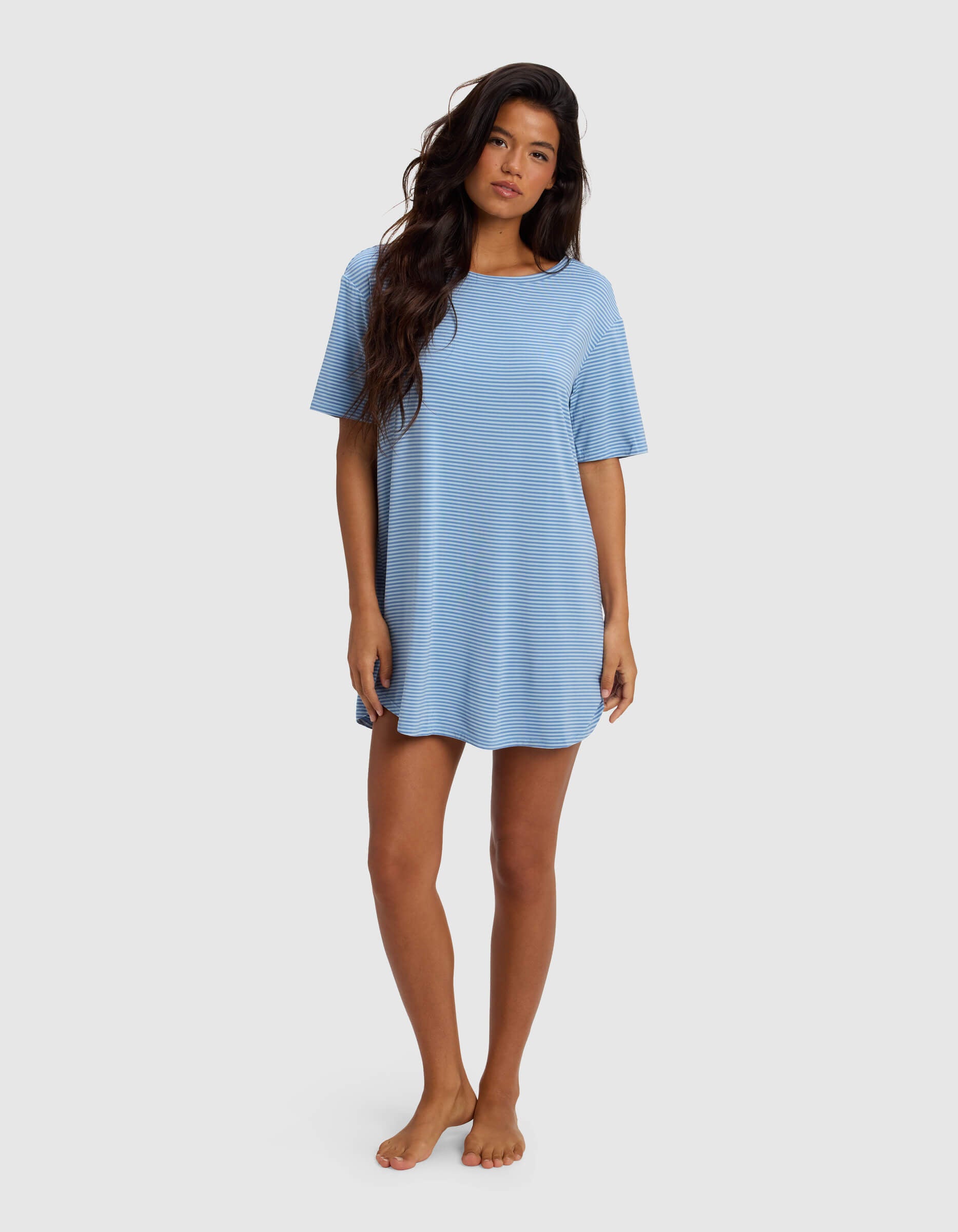 A woman with long wavy hair stands barefoot against a plain light background wearing the Cozy Earth Women's Bamboo Stretch Knit Sleep Dress, a loose, short-sleeve mini dress in light blue stripes.|Color:Sailor Mini Stripe
