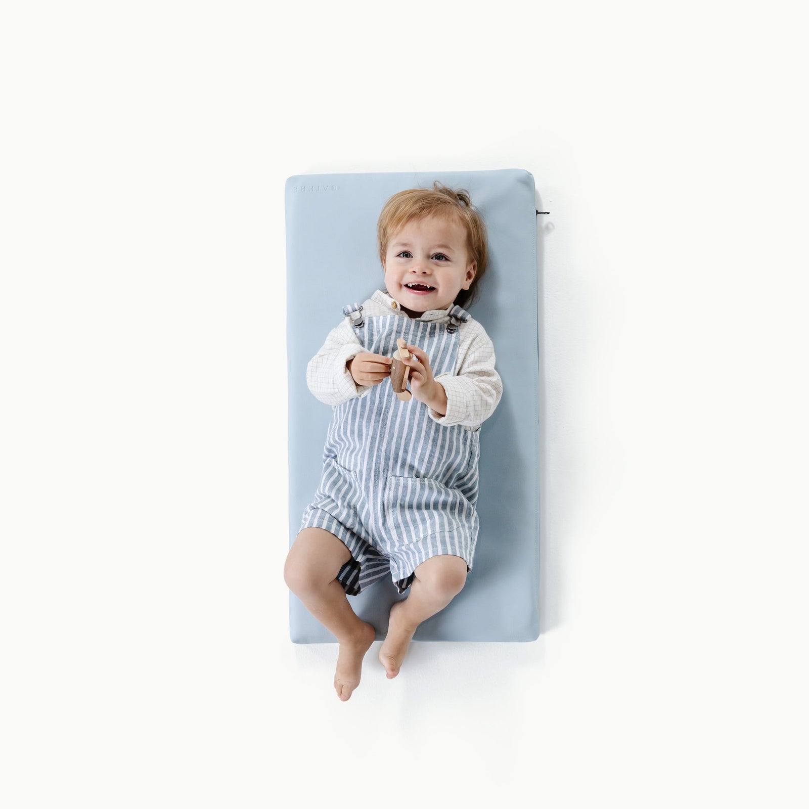 A smiling toddler lies on the Gathre Wholesale Padded Changing Mat, featuring a light blue color, with a white background.