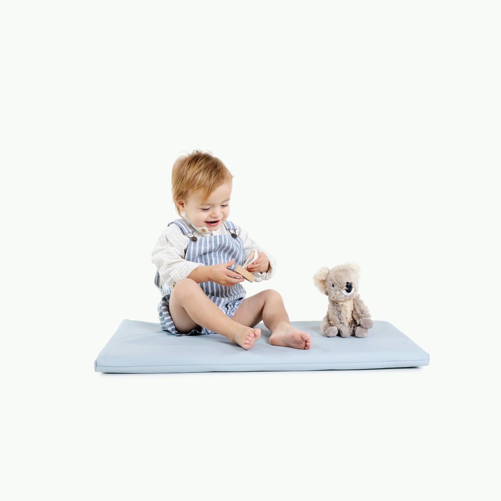 A smiling baby sits on the Gathre Wholesale Padded Changing Mat in Beau color, next to a plush teddy bear, against a plain white background.