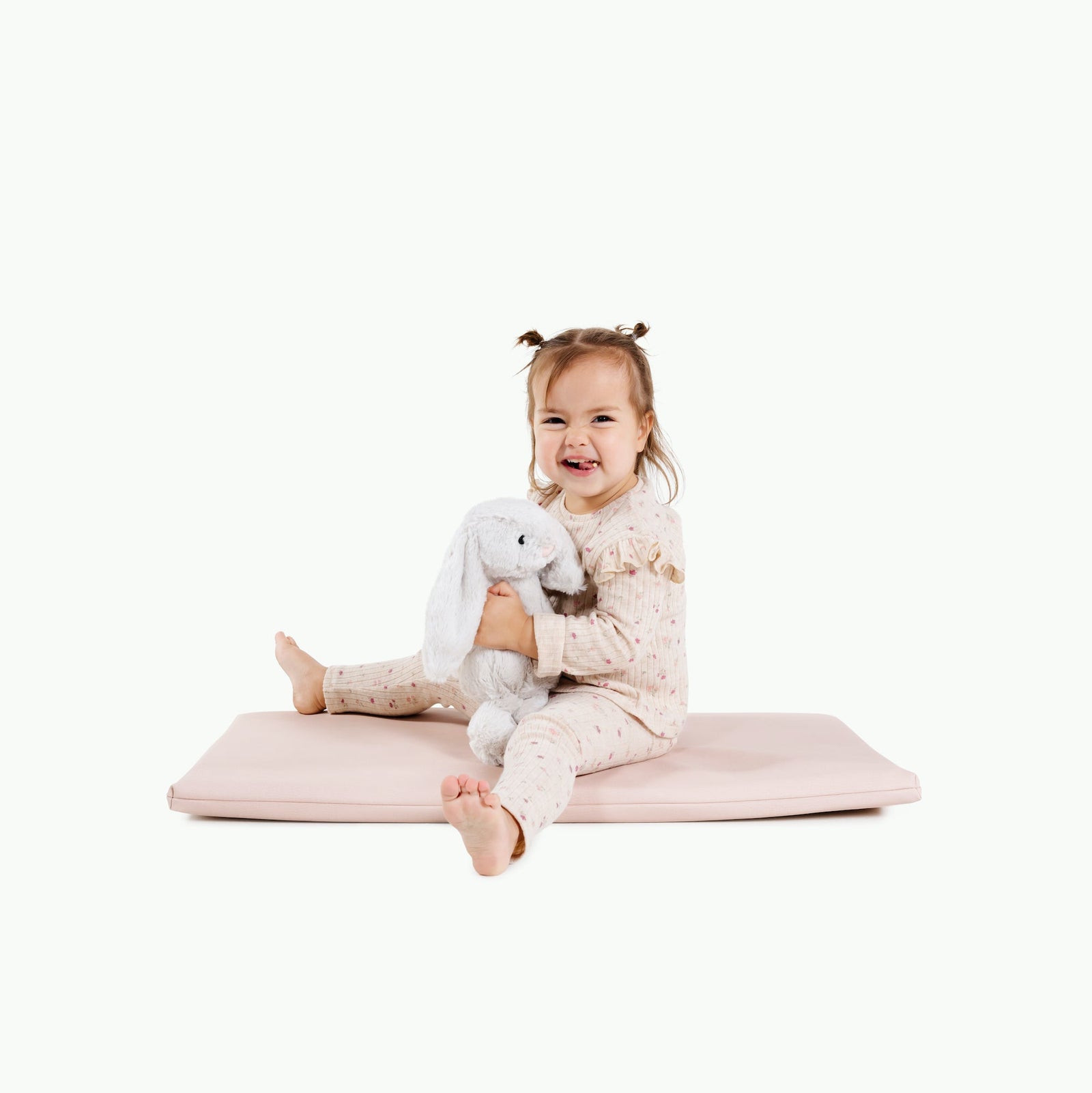A smiling toddler with pigtails sits on the Gathre Wholesale Padded Changing Mat in Belle, wearing light-print pajamas and hugging a soft gray bunny plush. The background is plain white.