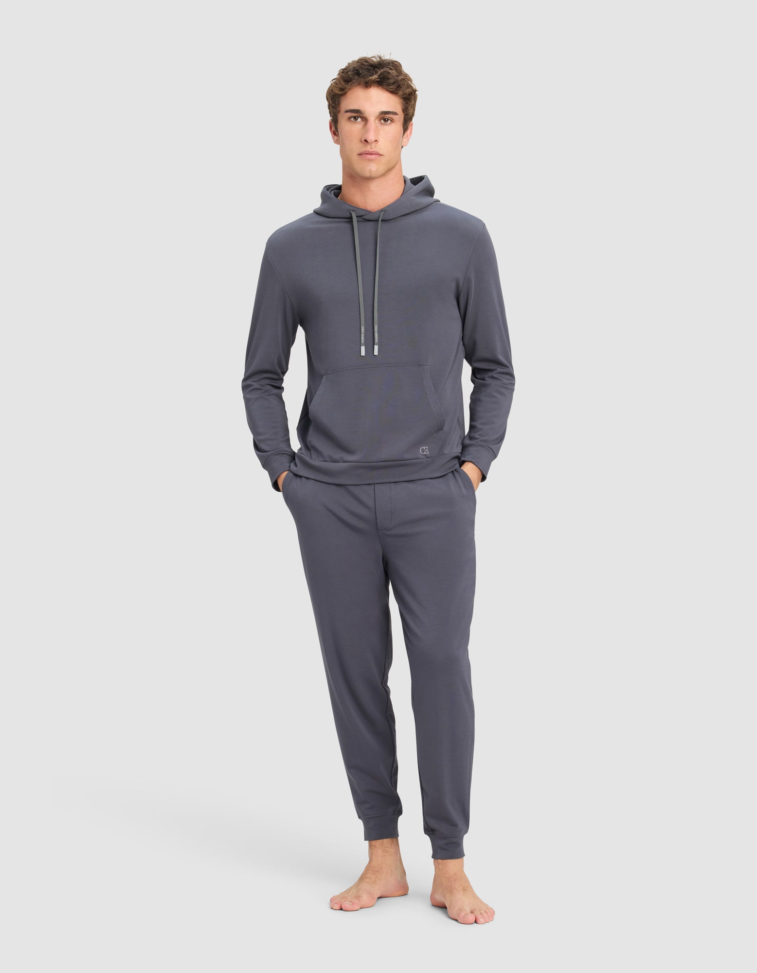 A young man stands barefoot against a plain background, hands in pockets, wearing the Cozy Earth Men's Brushed Bamboo Hoodie & Jogger Set in dark gray, and looks directly at the camera.