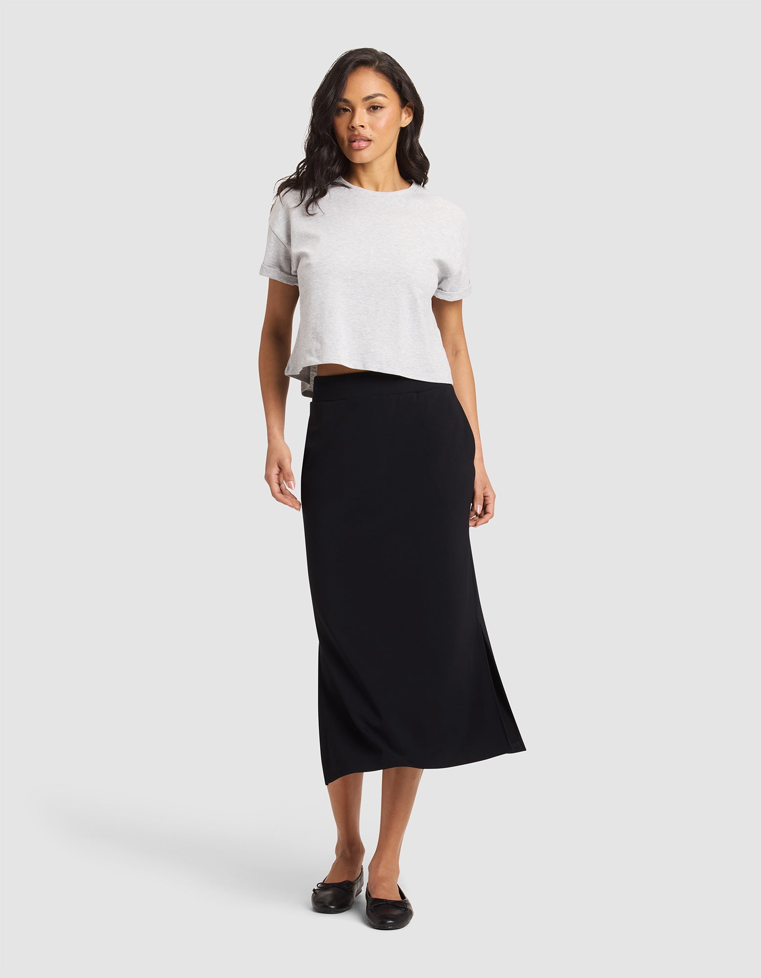A woman with wavy dark hair and a confident stance wears the Cozy Earth Women's Brushed Bamboo Midi Skirt in black, paired with a light gray short-sleeve top and black flats, standing against a plain background. |Color:Black