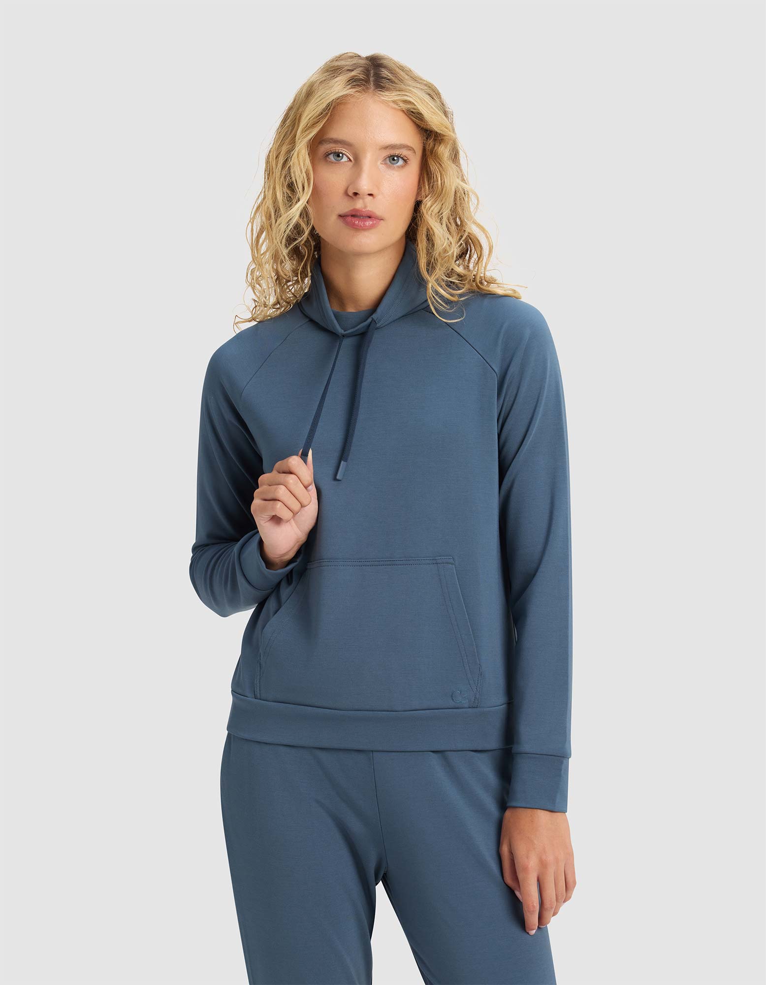 A woman with wavy blonde hair models the Cozy Earth Women's Brushed Bamboo Mock Neck in blue, standing against a plain light background. She holds the hoodie drawstring in one hand, wearing matching pants and a neutral expression.|Color:Dawn
